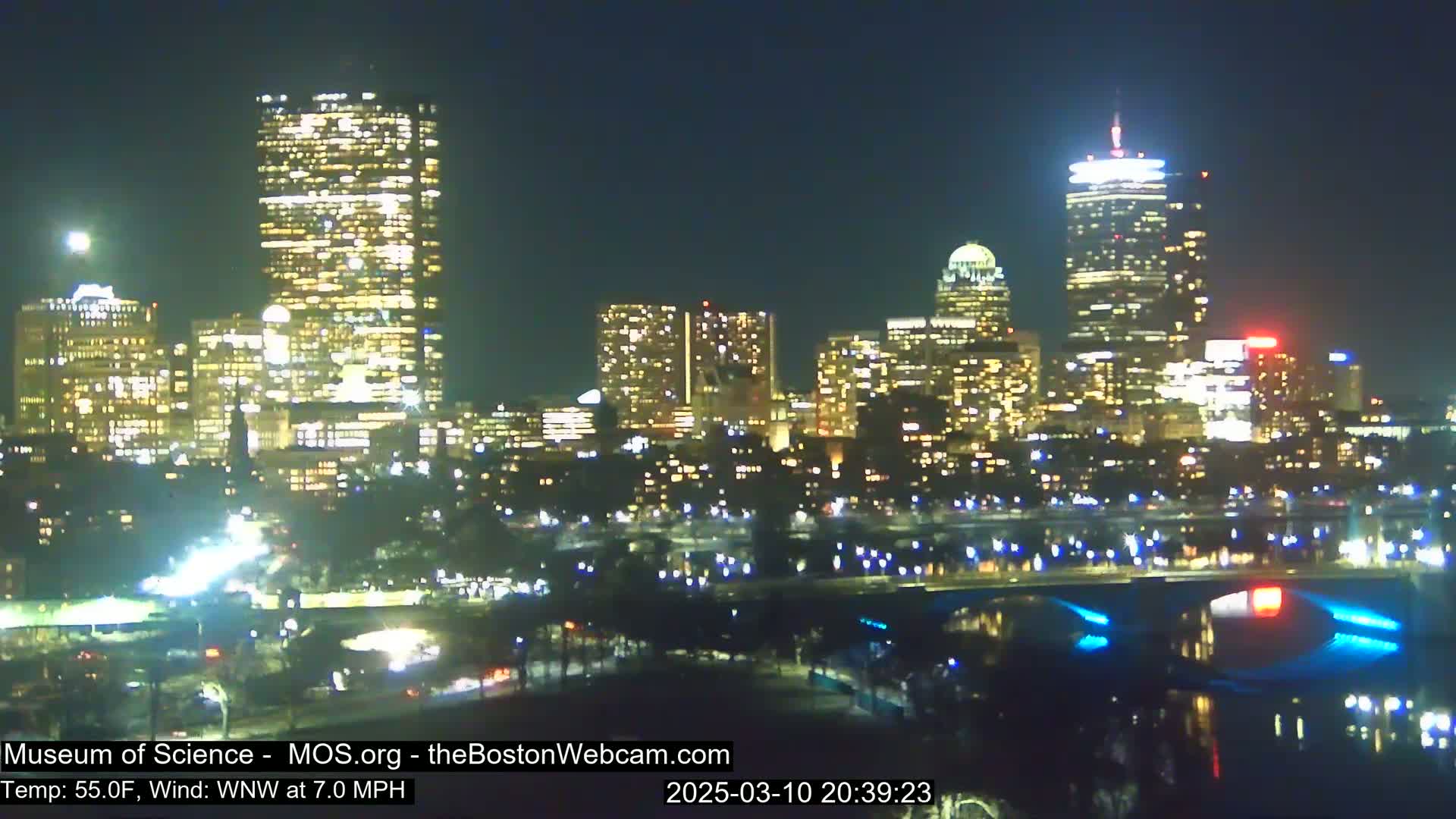 A nighttime view of a city skyline illuminated by many lights, with a temperature of 55°F and a west-northwest wind at 7.0 mph.