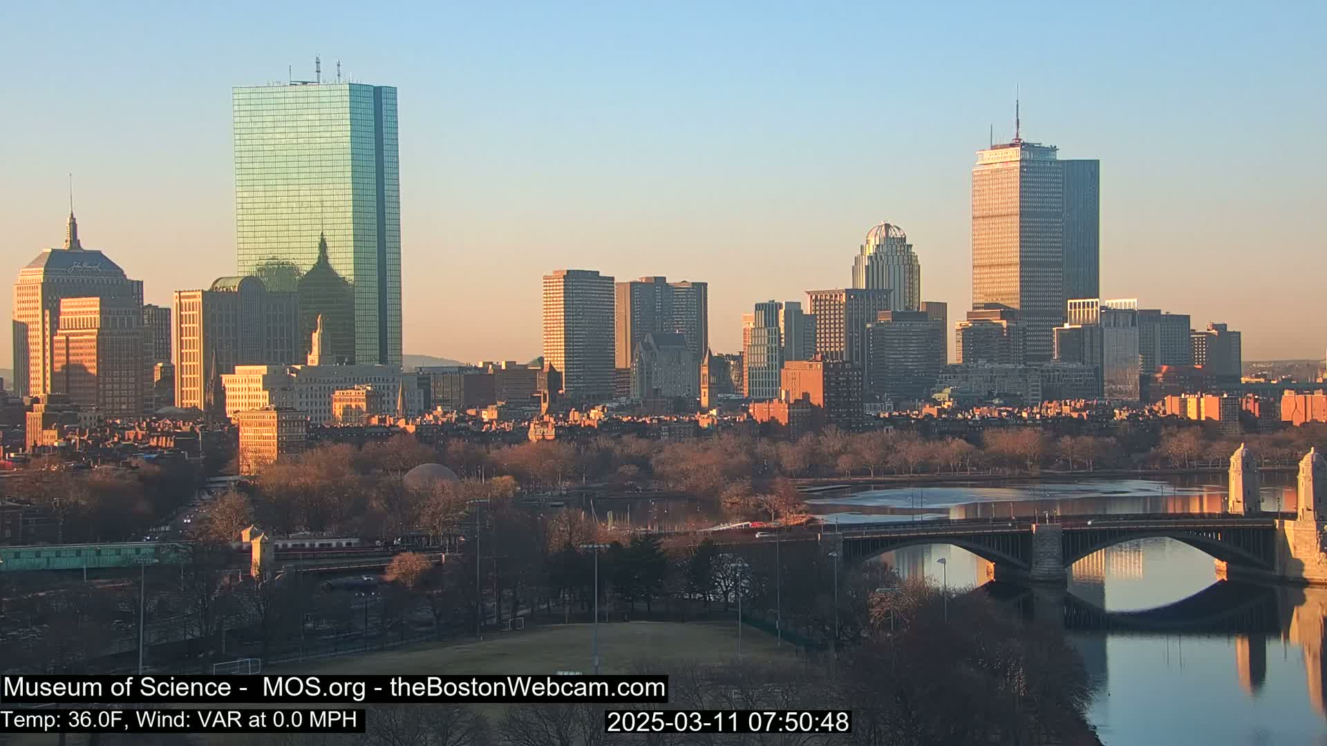 A city skyline at sunrise, with a bridge spanning a river in the foreground and a calm, clear sky.