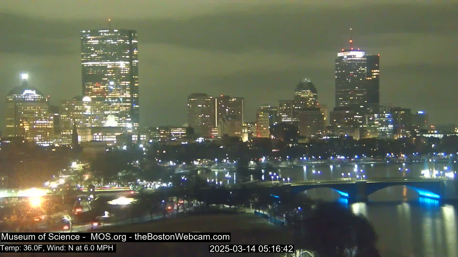 A nighttime cityscape with illuminated buildings across a river, under an overcast sky.