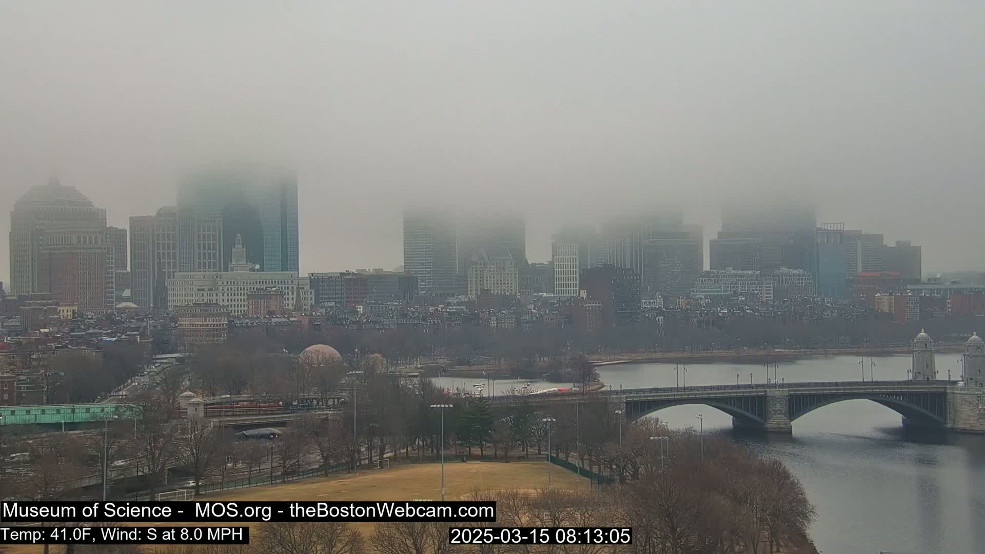 A foggy city skyline, partially obscured by mist, is seen across a river with a bridge in the foreground.