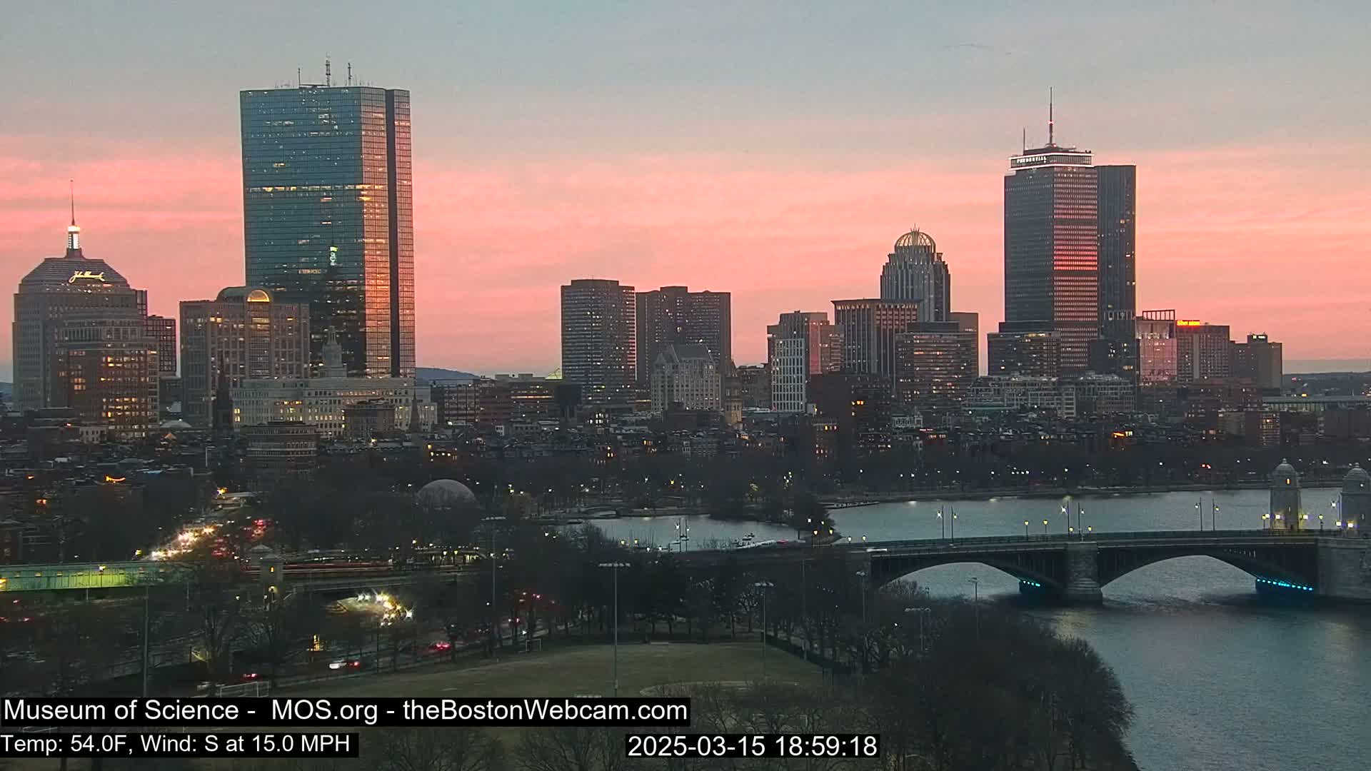 A city skyline at sunset with a pink sky, a river, and a bridge.