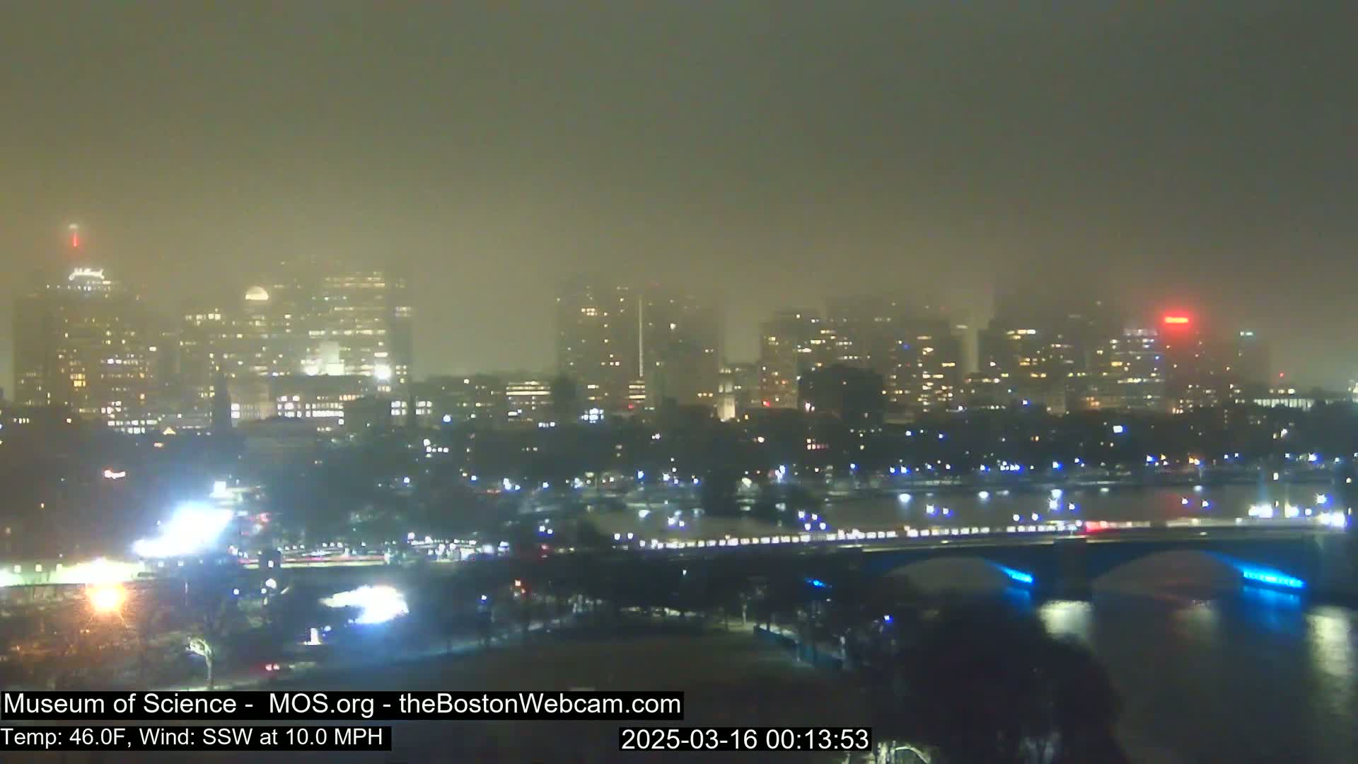 A hazy, nighttime view of a city skyline across a river, with a bridge illuminated in blue.