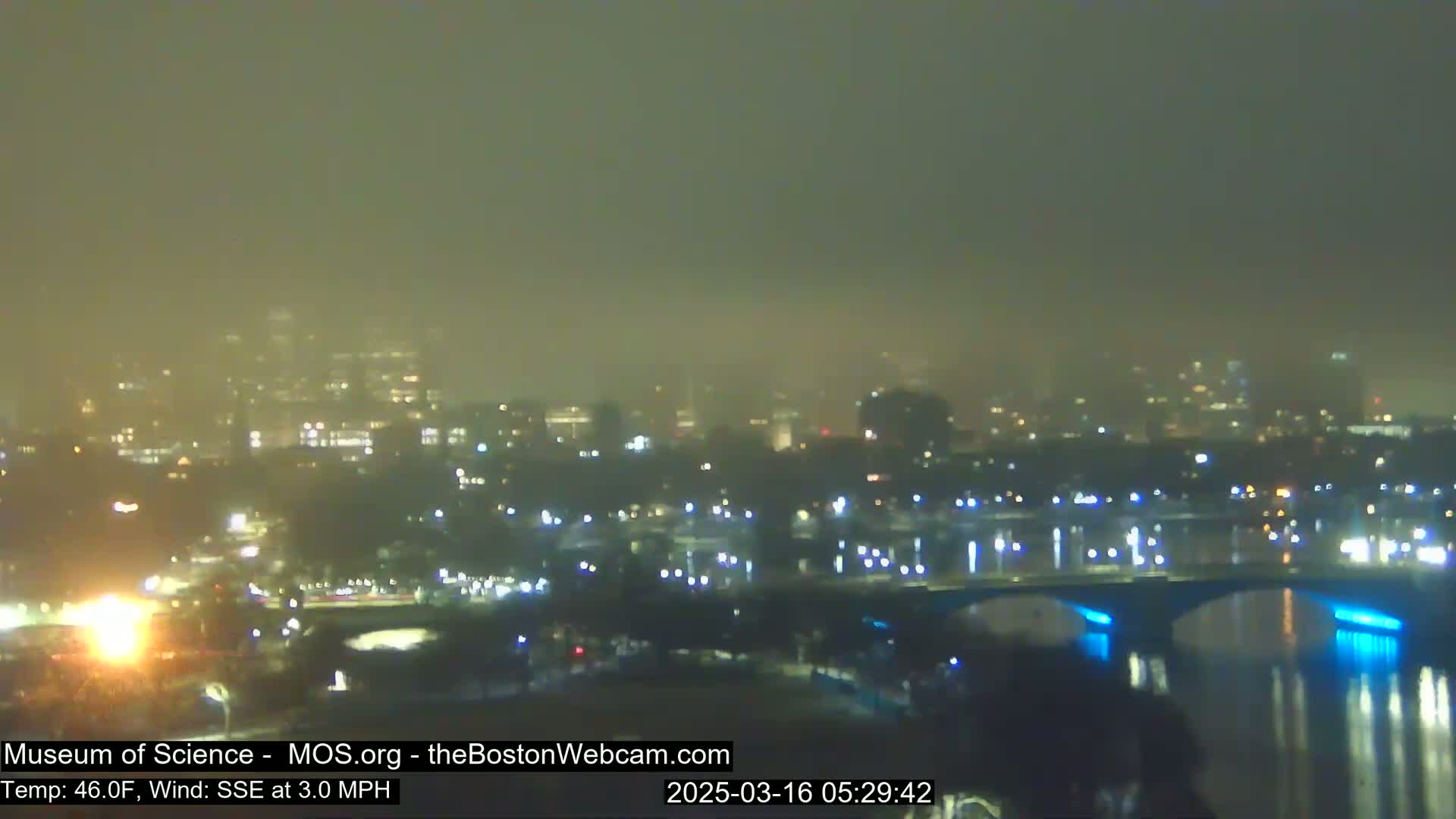 A hazy, nighttime view of a city skyline across a river with a bridge illuminated in blue.
