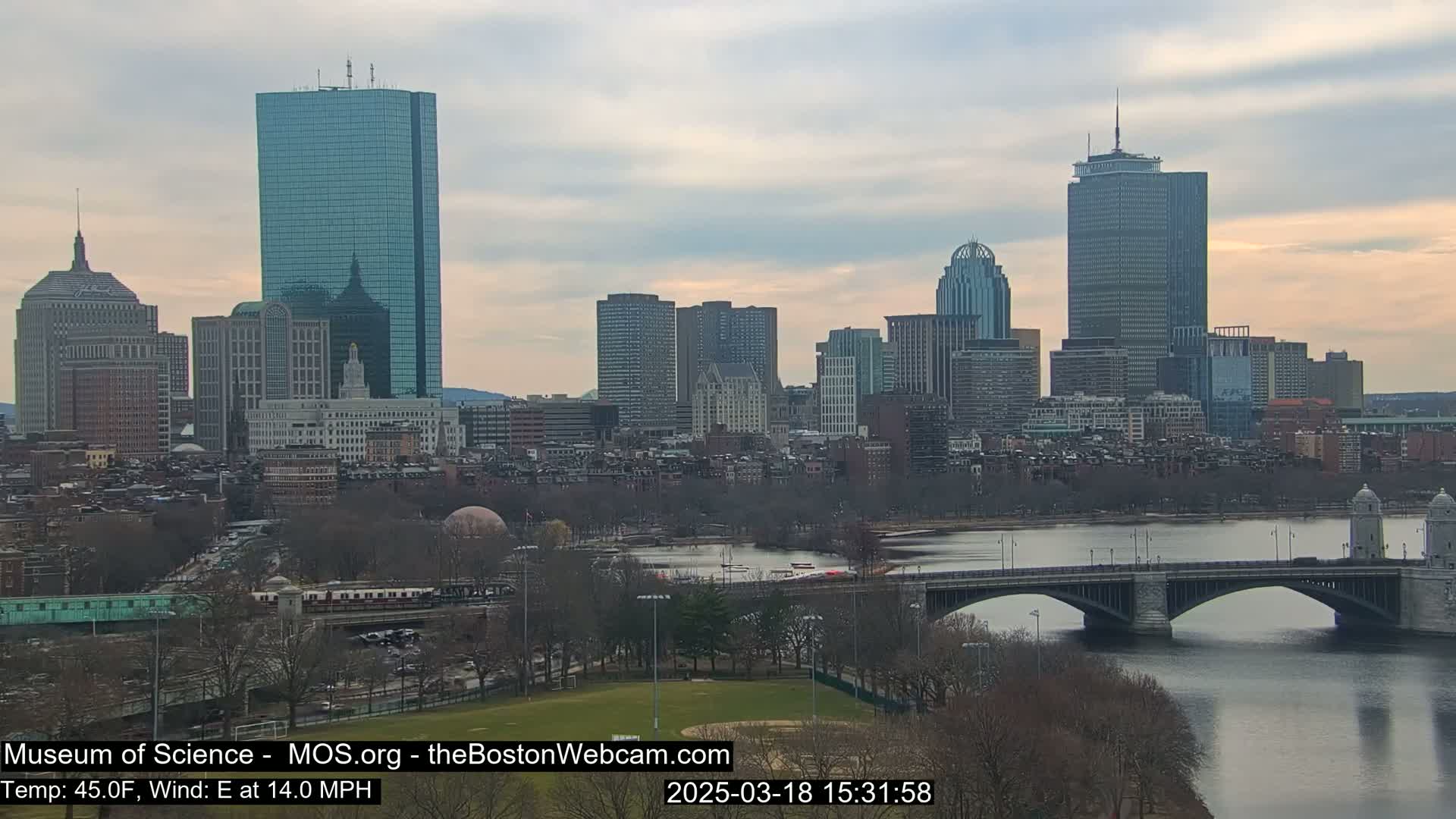 A city skyline with numerous skyscrapers and buildings is visible across a calm river with a bridge, under a cloudy, overcast sky.