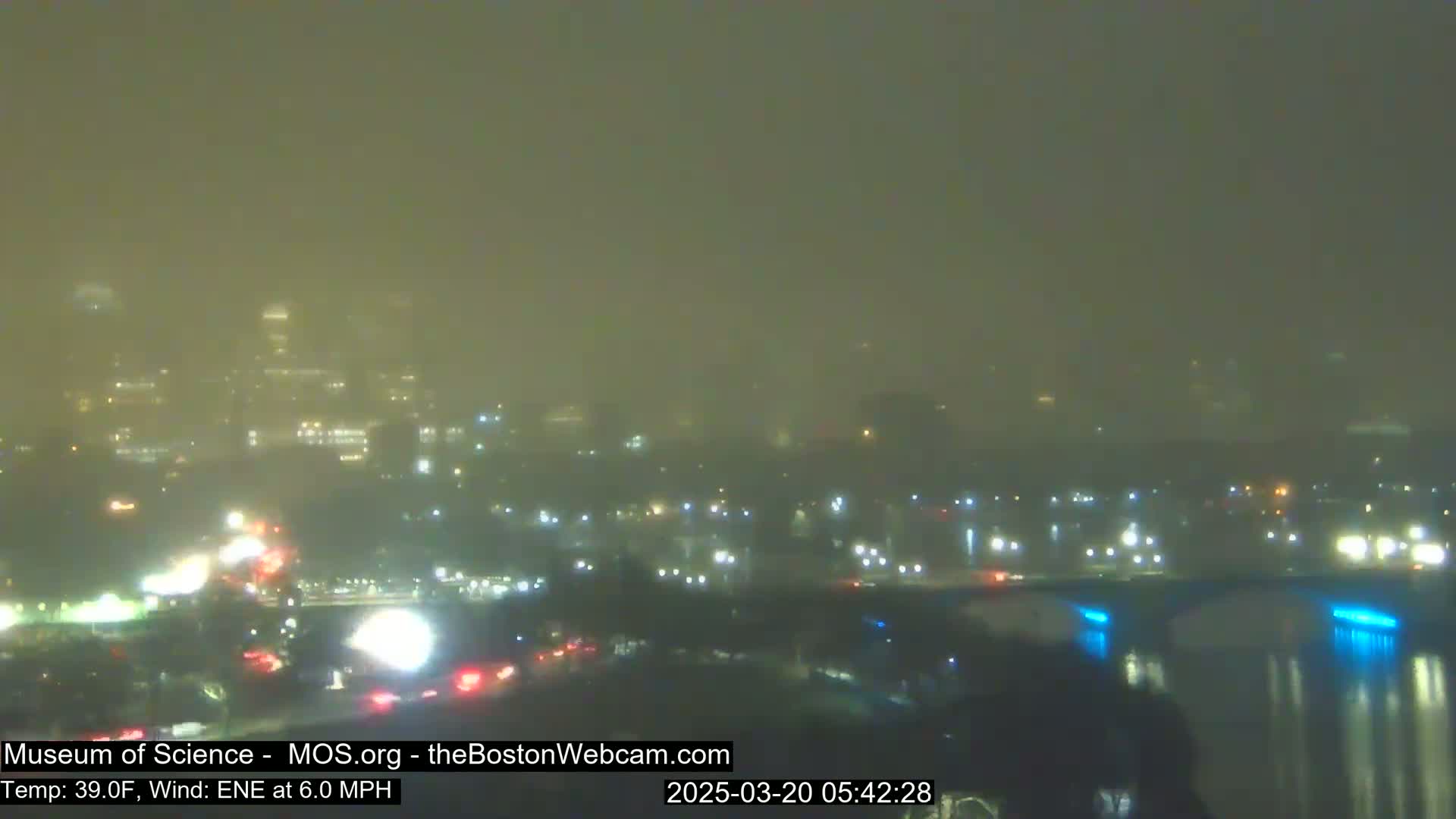 A hazy, night-time view of a city skyline and a highway with traffic, under a 39°F temperature and a 6.0 mph east-northeast wind.