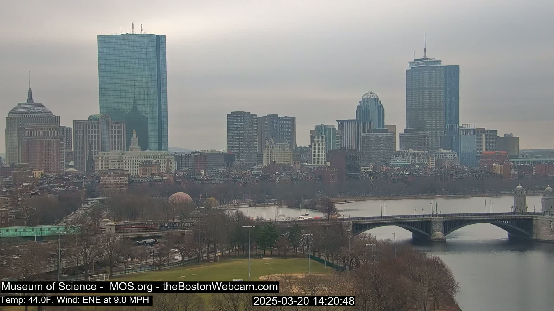 A cityscape with several skyscrapers is seen across a river and bridge under an overcast sky.