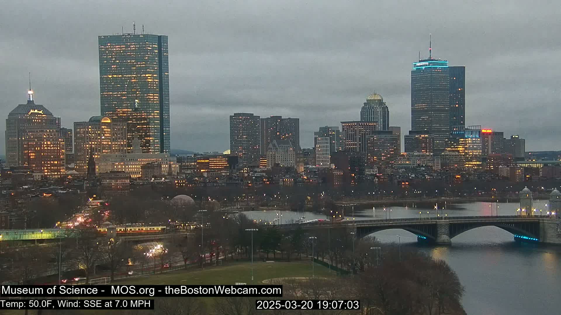 A nighttime cityscape with several illuminated skyscrapers, a river, and a bridge under an overcast sky.