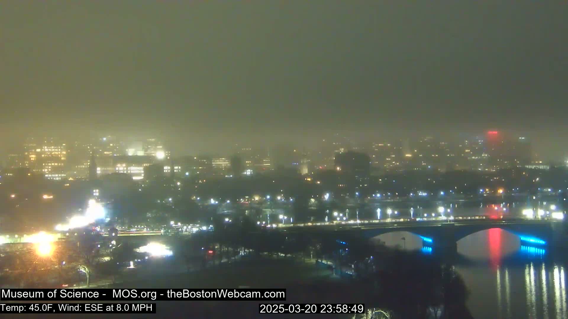 A foggy night scene shows a city skyline with illuminated buildings and a bridge over a body of water.