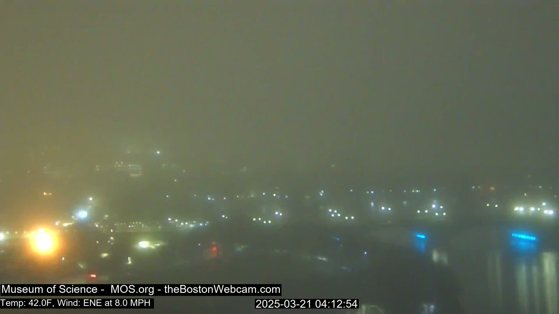 A foggy, nighttime scene shows a dimly lit cityscape with scattered lights visible through the haze.