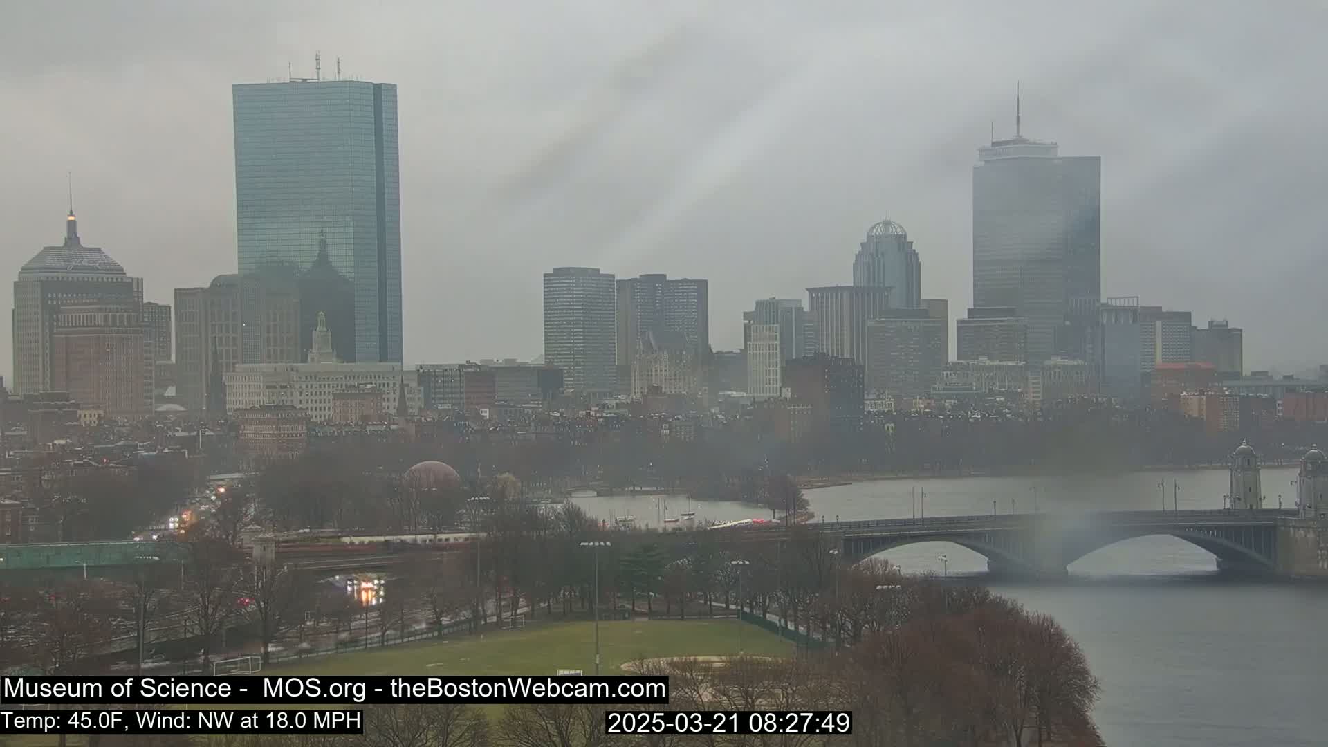 A hazy, overcast cityscape with several tall buildings is visible across a river, with a bridge in the foreground and bare trees along the riverbanks.