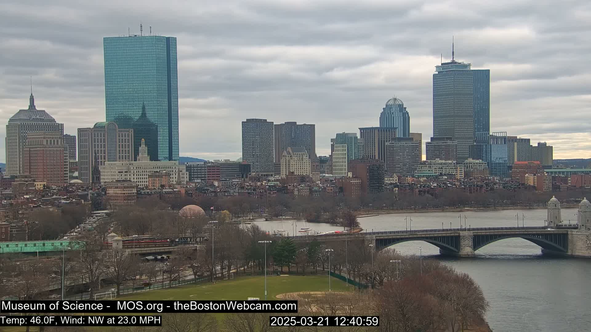 A cityscape with numerous skyscrapers overlooks a river and bridge under a cloudy sky.