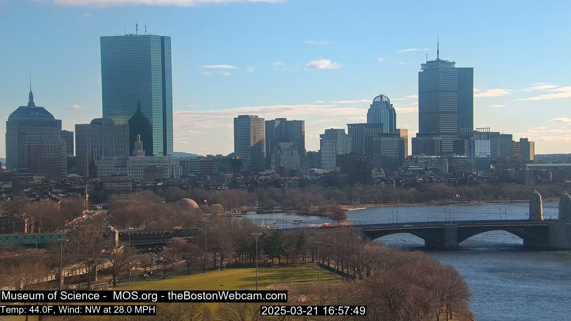 A city skyline with several skyscrapers is visible across a river, under a mostly clear, sunny sky; a bridge spans the river in the foreground.