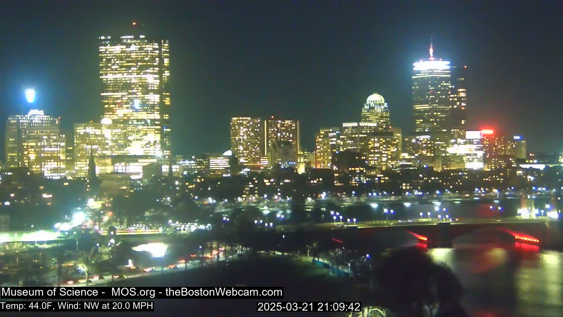 A nighttime cityscape illuminated with many bright lights, seen across a body of water with a bridge, under a clear sky with a northwest wind at 20 mph and a temperature of 44°F.