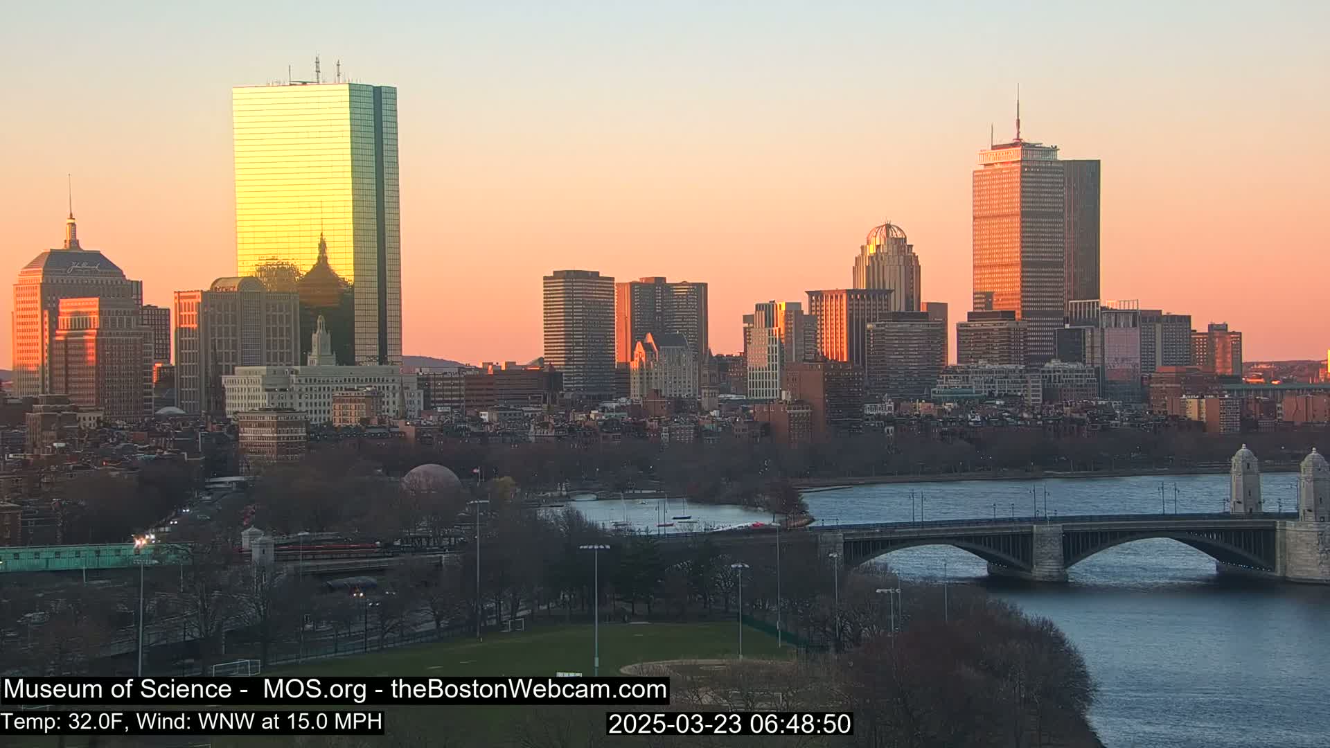 A city skyline at sunset, with a bridge crossing a river in the foreground, under a clear, orange-hued sky.
