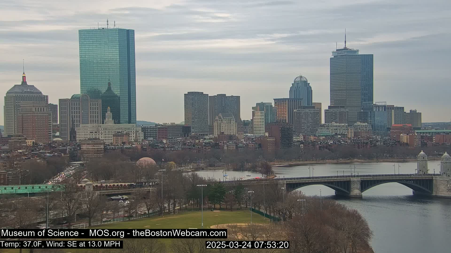 A city skyline with several tall buildings is visible across a river, under an overcast sky.