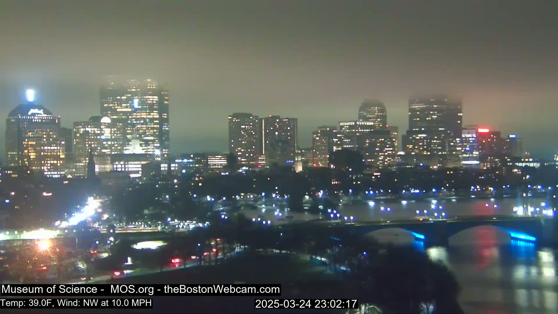 A nighttime cityscape shrouded in fog, with a bridge spanning a body of water in the foreground.
