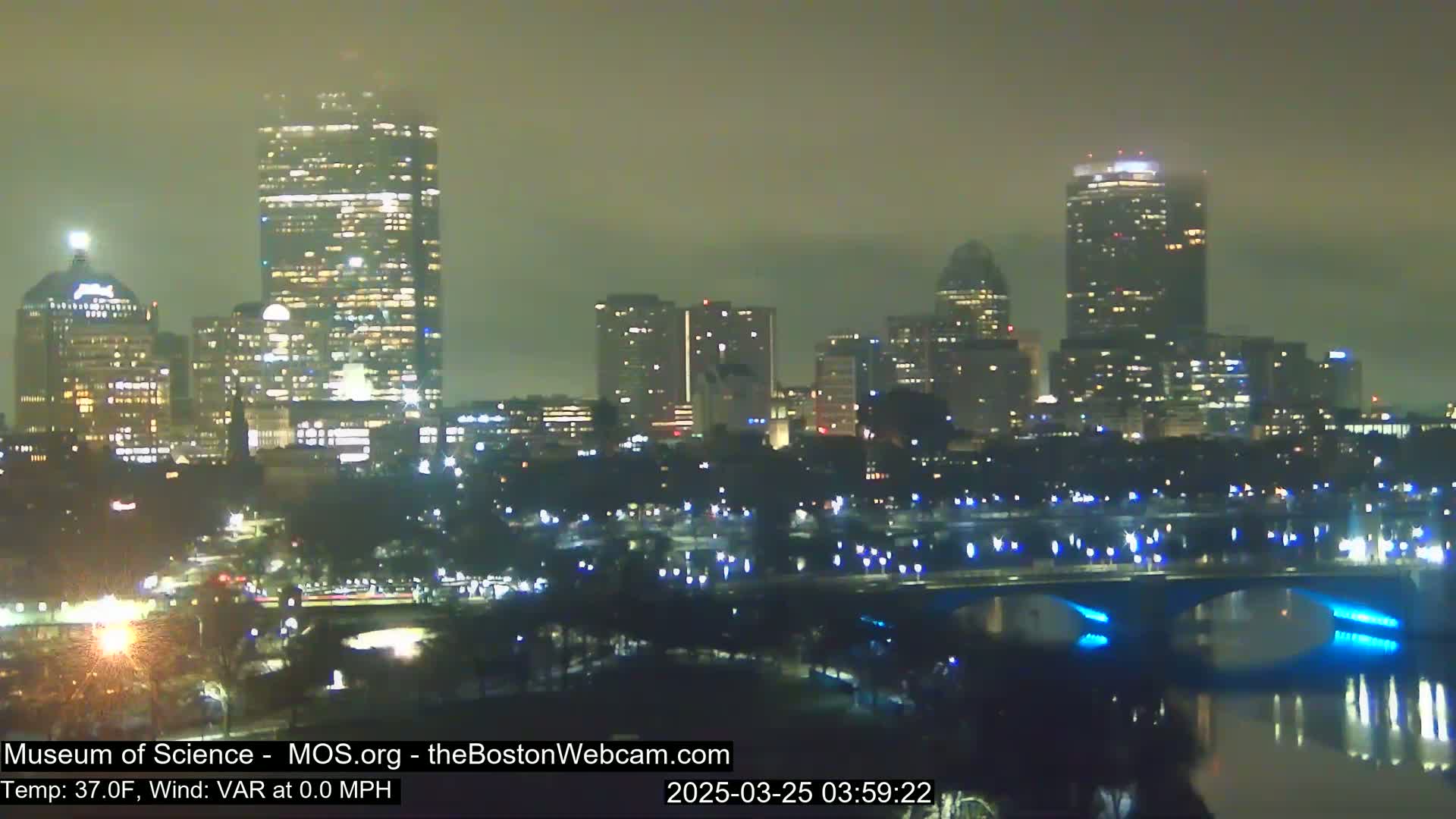 A nighttime cityscape, shrouded in fog, shows numerous illuminated buildings along a river with a bridge displaying blue lights.