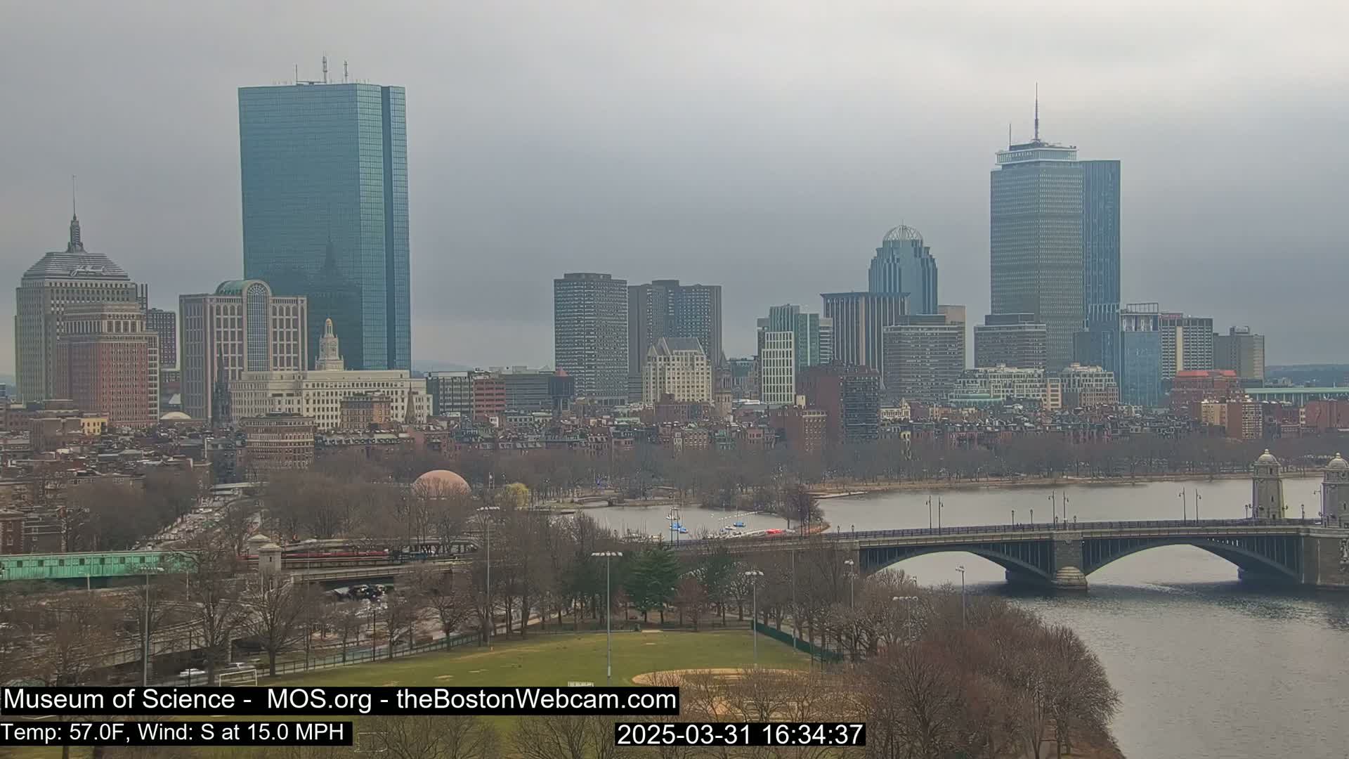 A cityscape with several skyscrapers is visible across a river, with a bridge and a park in the foreground, under an overcast sky.