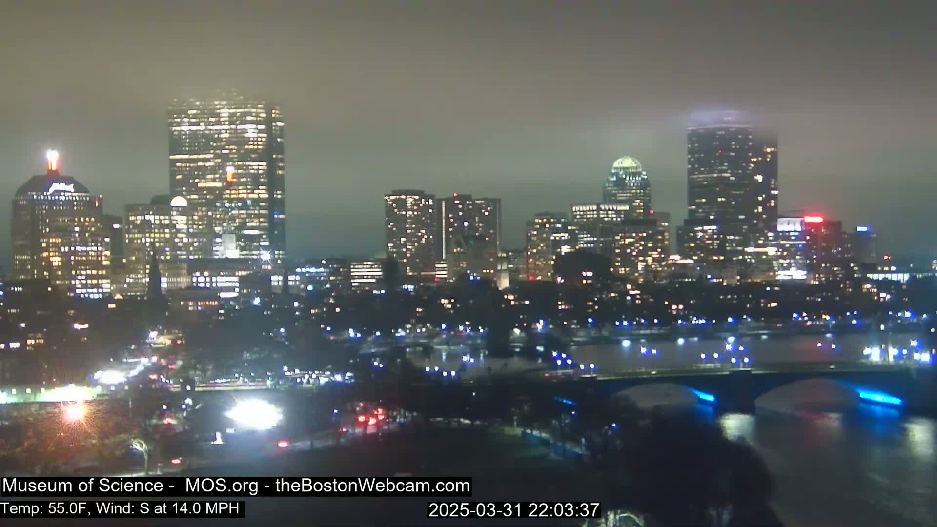 A nighttime cityscape with numerous illuminated buildings is visible across a body of water, under hazy conditions with a south wind.