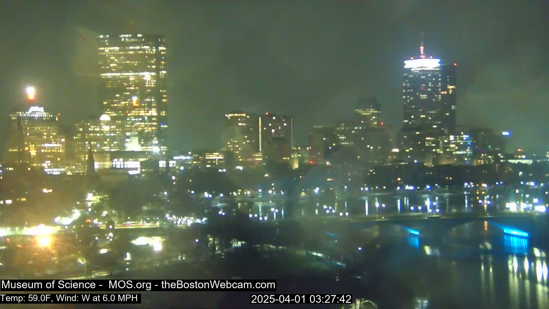 A nighttime view of a city skyline across a body of water, illuminated by city lights under slightly overcast conditions.