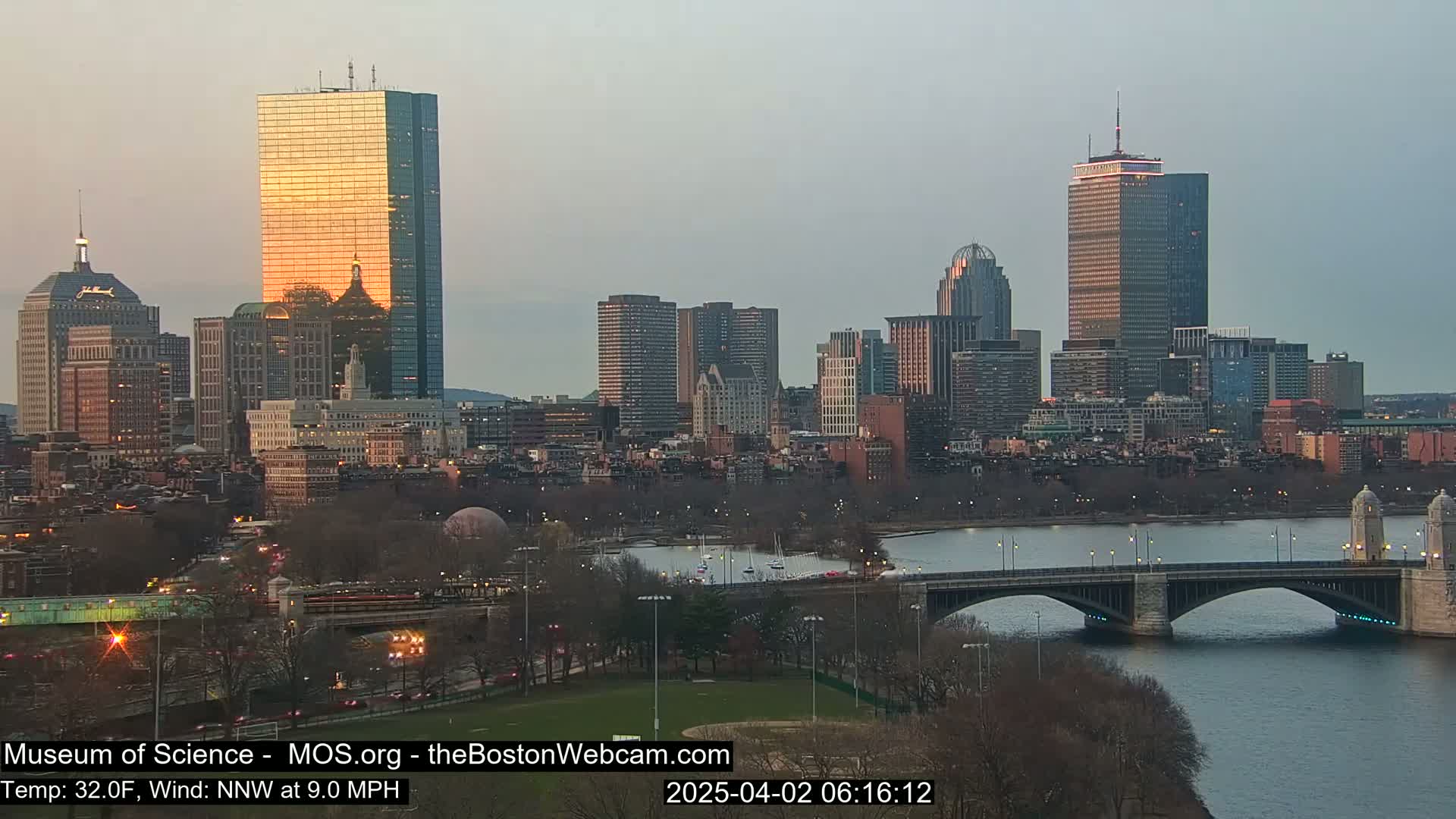 A city skyline at sunset, with a bridge spanning a river in the foreground, under a mostly clear sky.