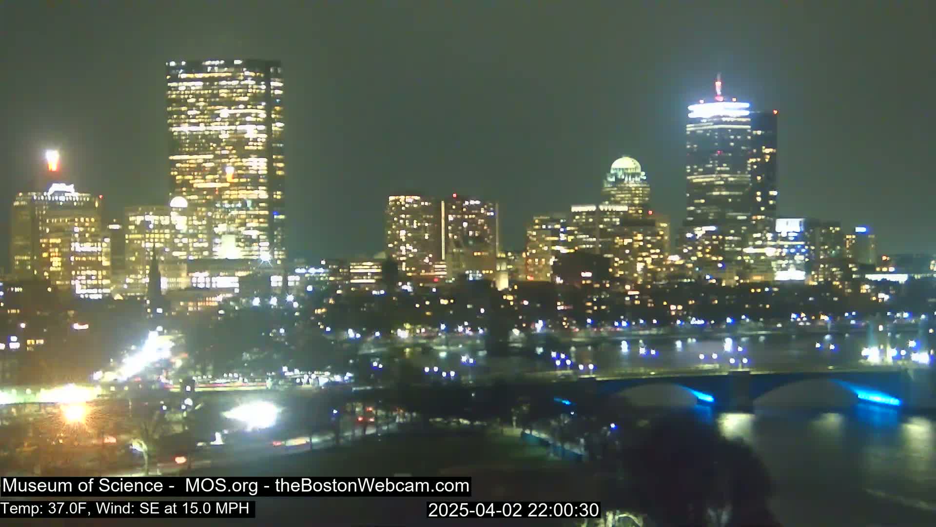 A nighttime view of a city skyline with brightly lit skyscrapers and bridges across a body of water, with a southeast wind at 15 mph and a temperature of 37 degrees Fahrenheit.