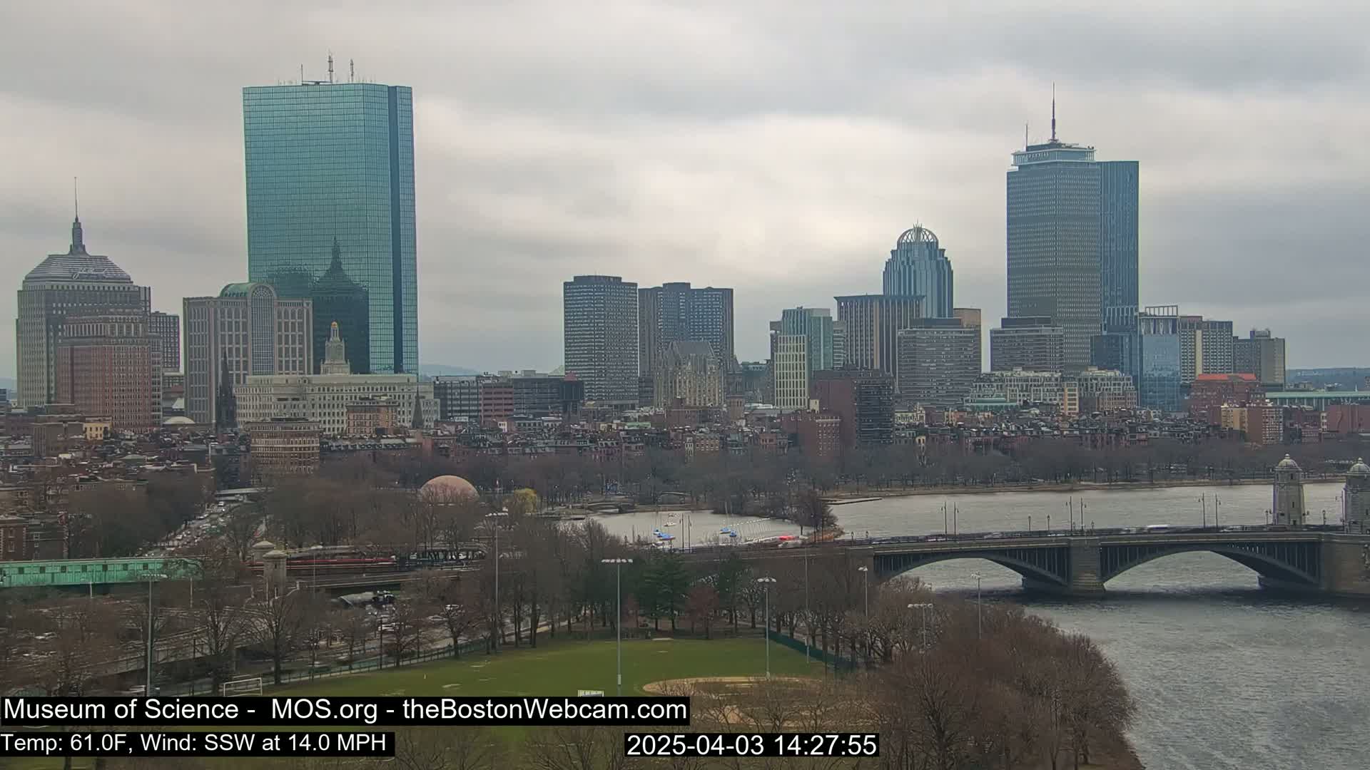 A cityscape with several skyscrapers is seen across a river and bridge under an overcast sky.