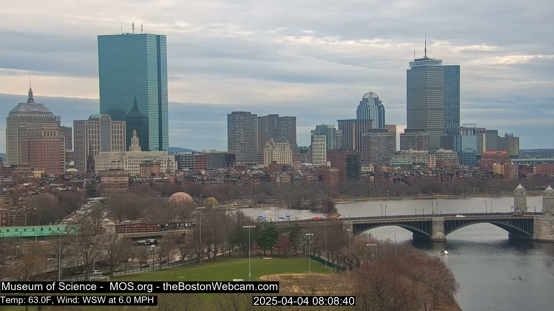 A cityscape with numerous skyscrapers and other buildings is viewed from across a river, under a cloudy sky, with a bridge in the foreground.