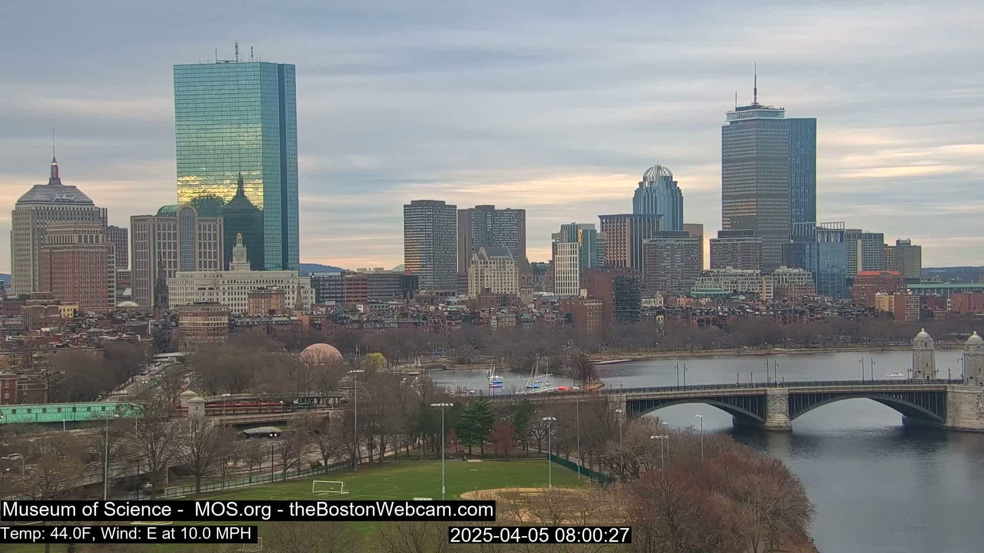 A city skyline with several tall buildings is seen across a river and bridge under a mostly cloudy sky.