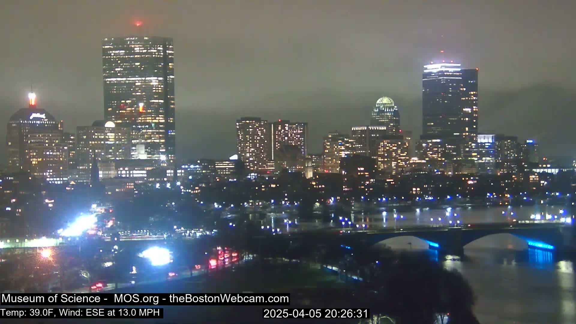 A nighttime cityscape with illuminated buildings and a bridge over a river under overcast skies, with a wind blowing from the east-southeast at 13 miles per hour.