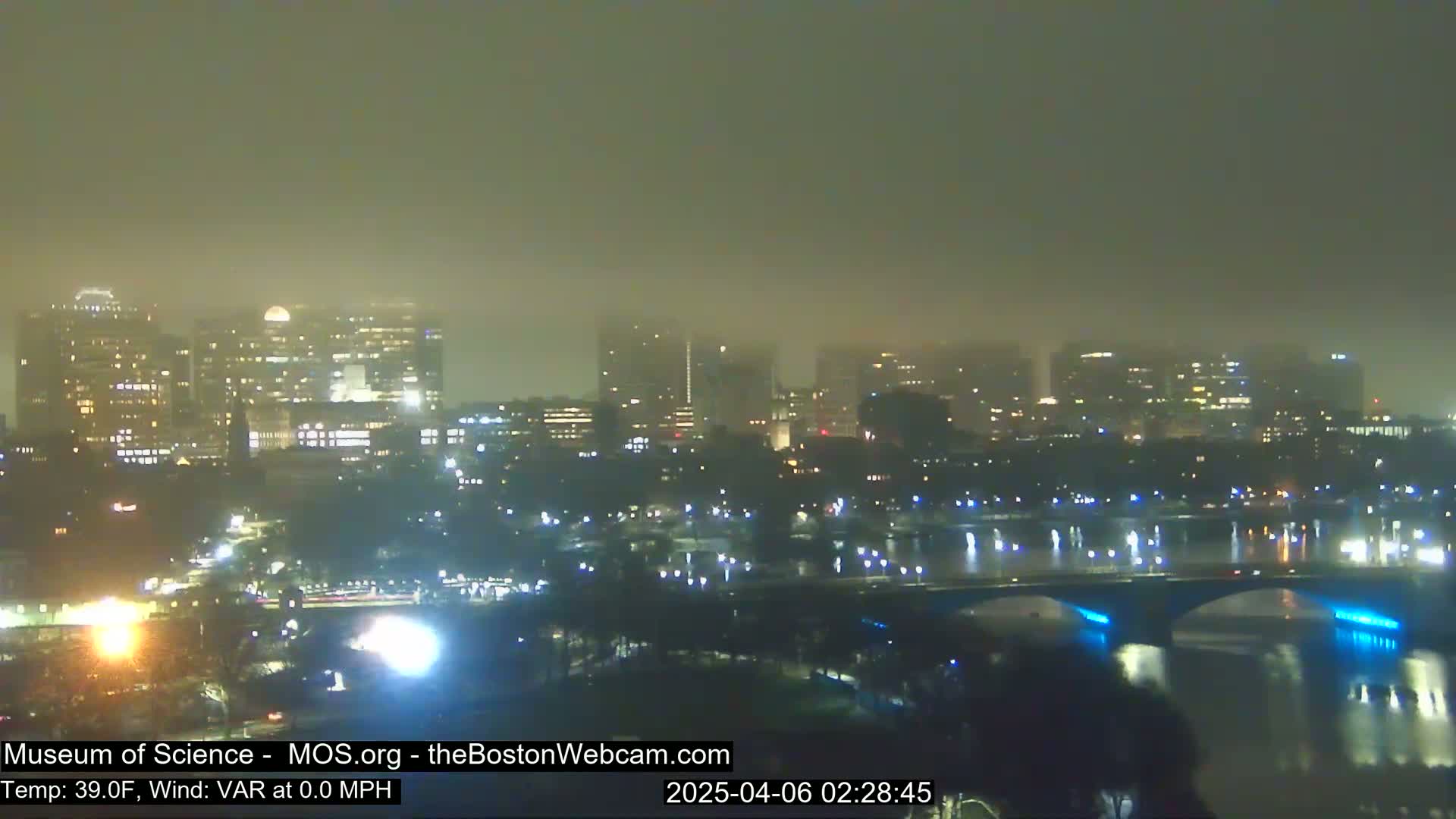 A foggy, nighttime cityscape with a river and bridge, illuminated by city lights.