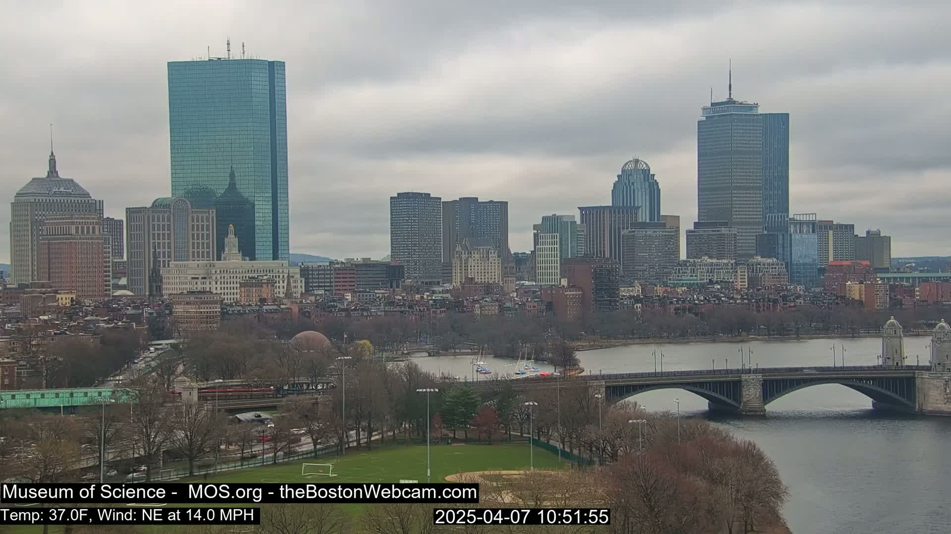 A city skyline with numerous skyscrapers is visible across a river and bridge under an overcast sky.