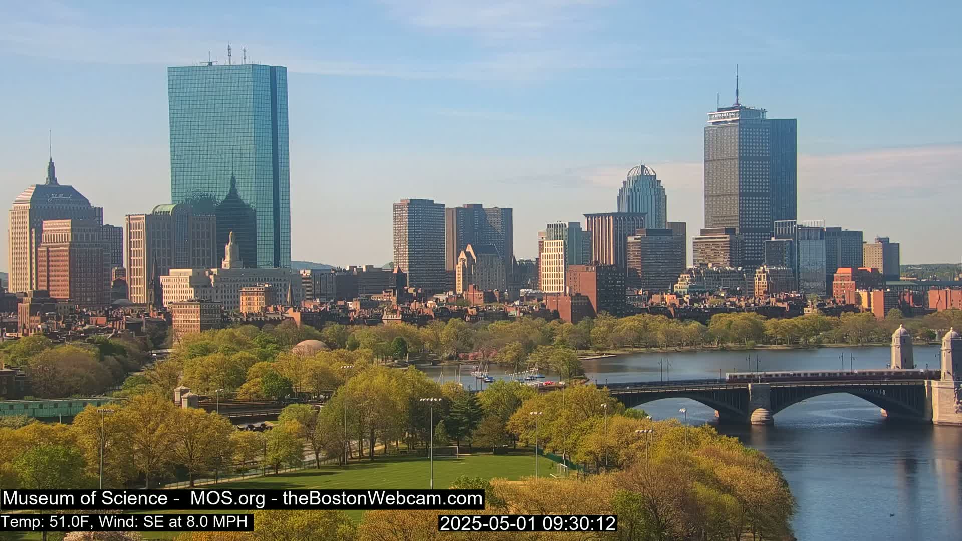 A sunny cityscape with a river, bridge, and green parkland in the foreground.