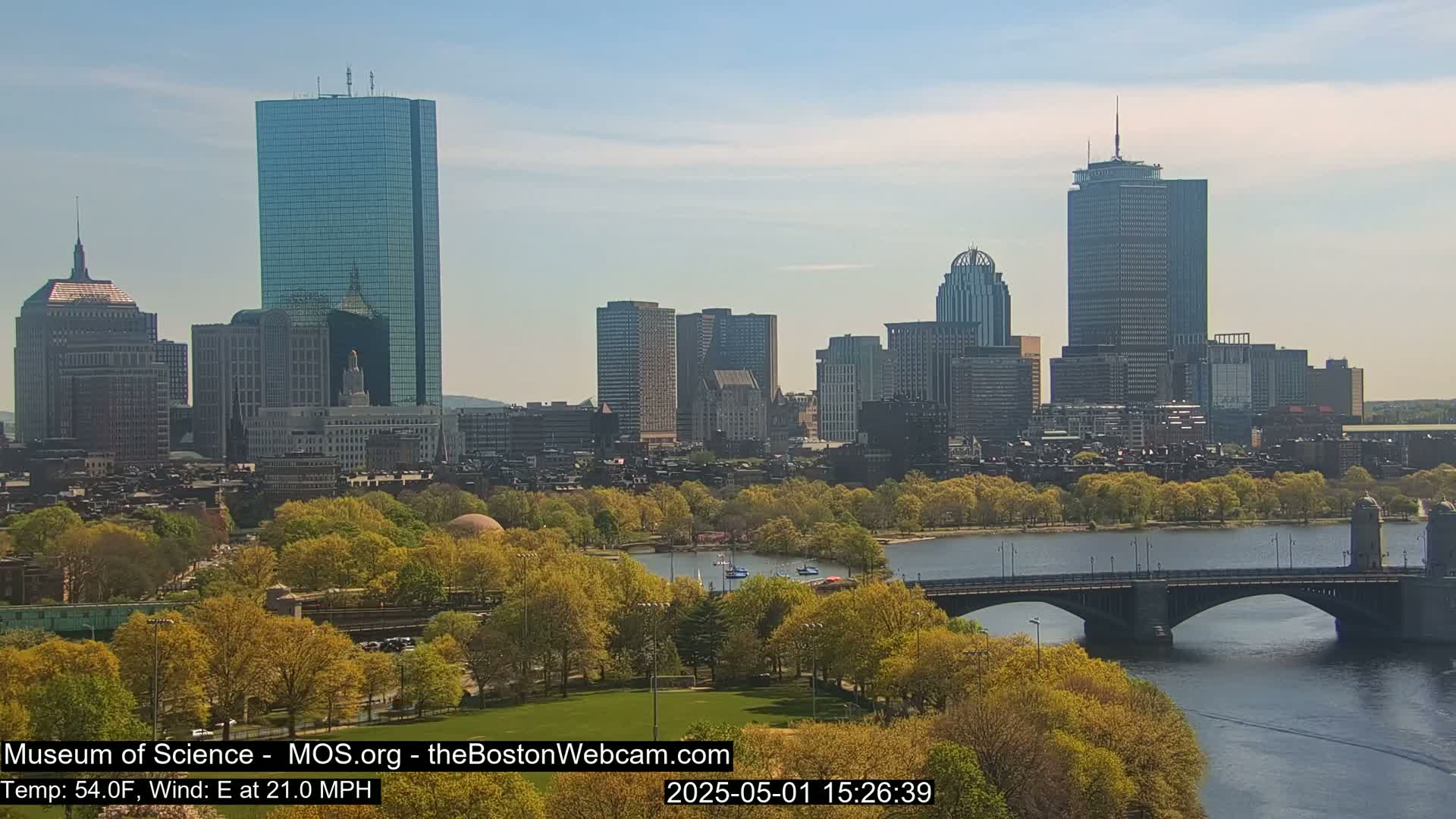 A cityscape with numerous skyscrapers is seen across a river with a bridge, under a partly sunny sky; trees are budding and in shades of green and yellow.
