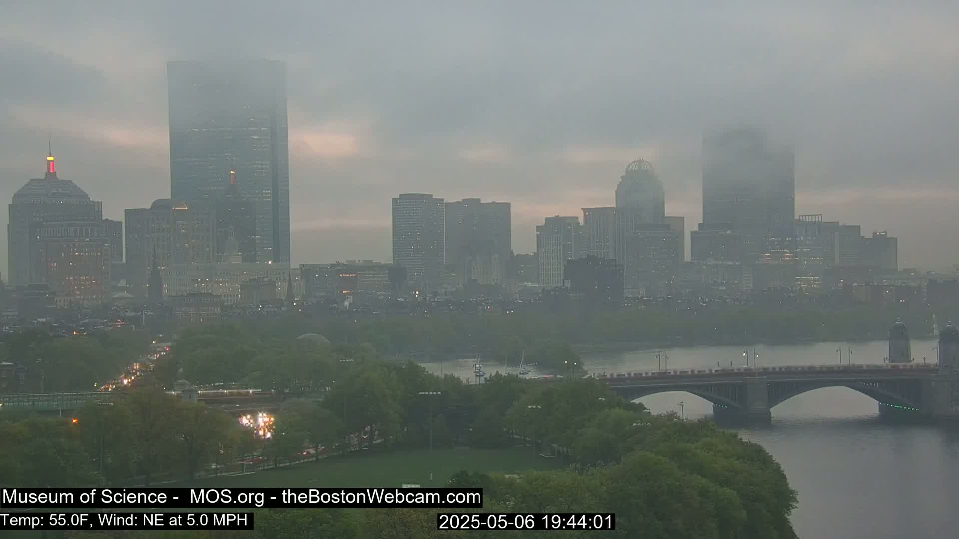 A foggy cityscape, shrouded in mist, shows a city skyline with a bridge over a river, and green trees along the riverbank.