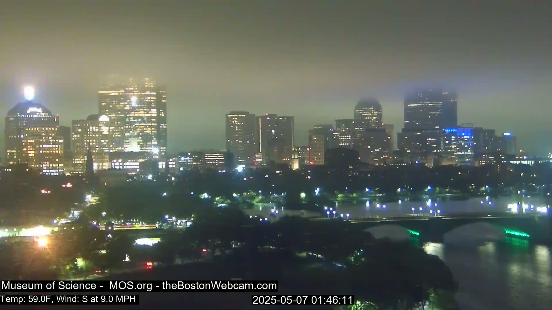 A nighttime view of a city skyline shrouded in fog, with a bridge and body of water in the foreground.