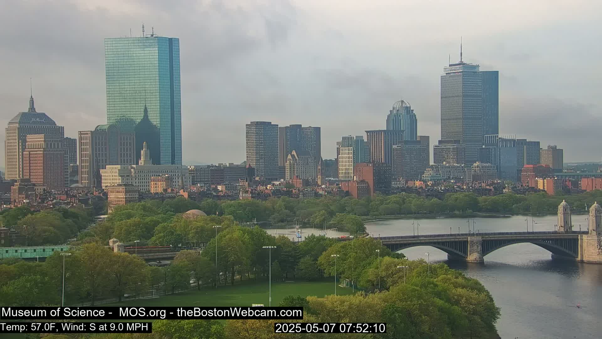 A cityscape with numerous skyscrapers and green parkland sits beside a river crossed by a bridge under a cloudy sky.