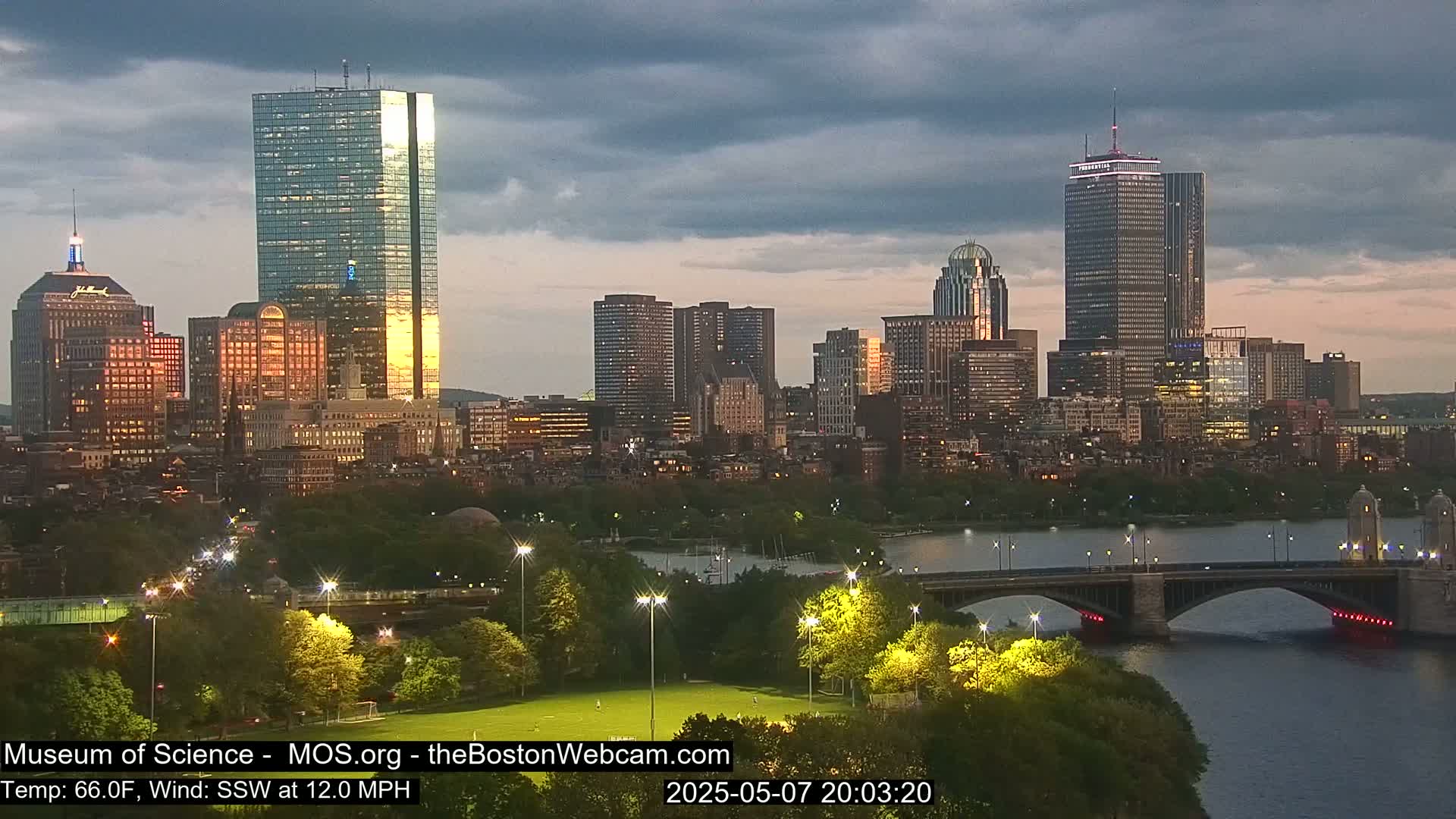 A city skyline at dusk under a cloudy sky, with a river and bridge in the foreground, and a park with lights.
