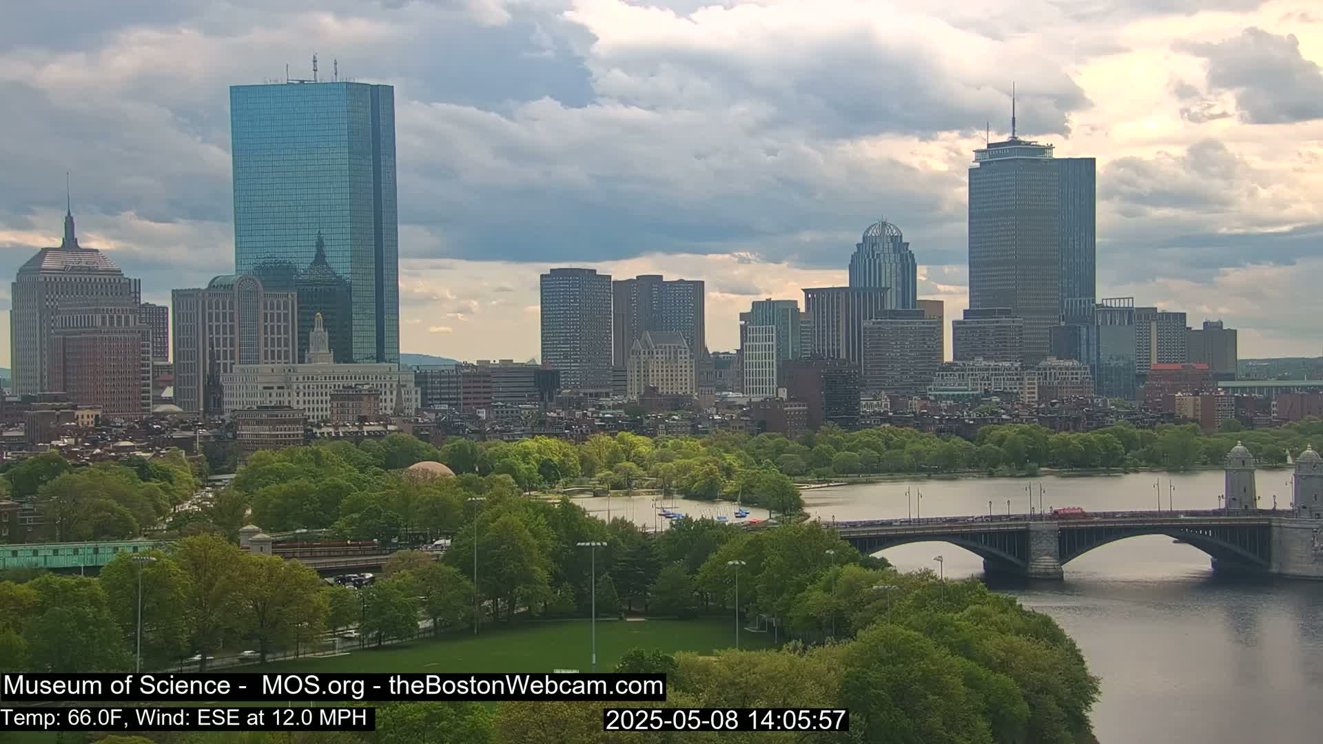 A cityscape with several skyscrapers is visible under a partly cloudy sky, with a bridge spanning a river in the foreground and lush green trees along the riverbank.
