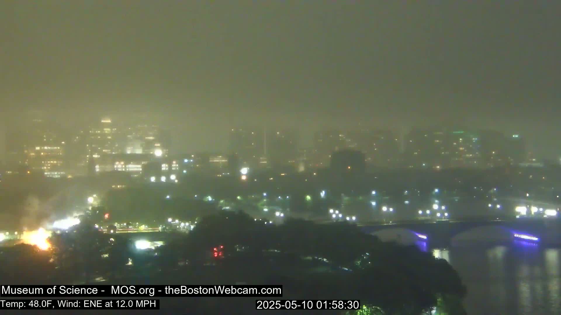 A foggy, nighttime cityscape is seen across a body of water, with a bridge and faint lights visible through the haze.