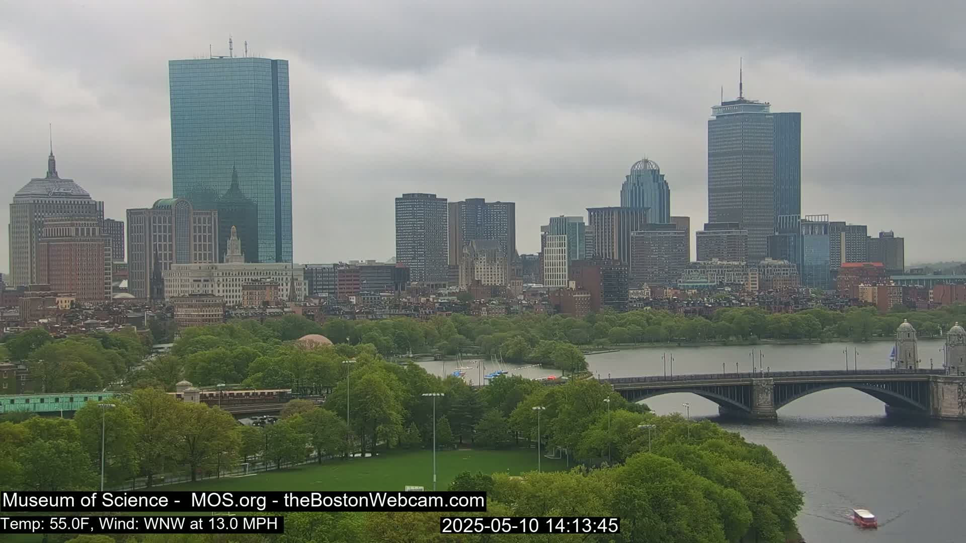 A city skyline with numerous buildings is visible across a river under a cloudy sky, with a bridge spanning the river and green parkland in the foreground.