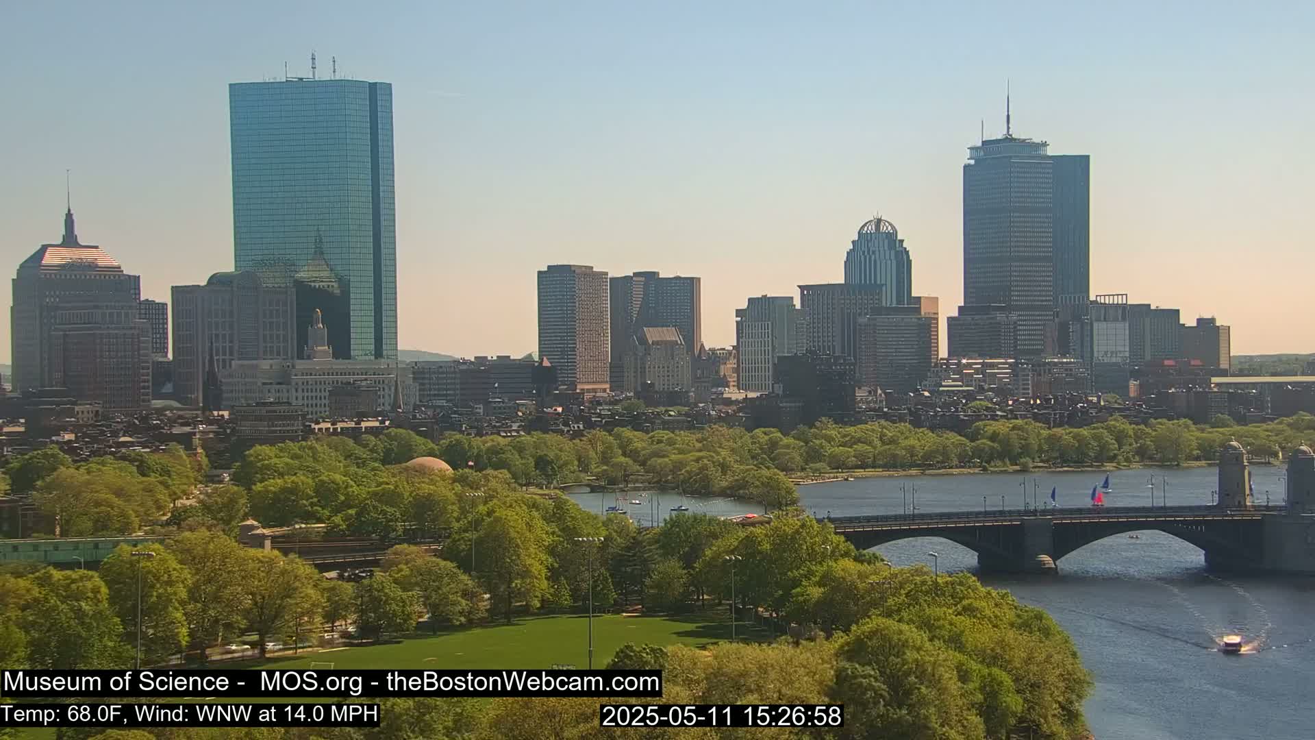 A sunny cityscape with numerous skyscrapers is seen across a river with a bridge, lush green trees, and a few boats.