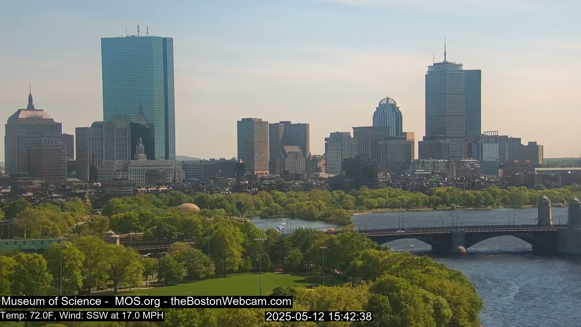 A sunny cityscape with numerous skyscrapers, green trees, and a bridge over a body of water, with a south-southwest wind at 17 mph and a temperature of 72°F.