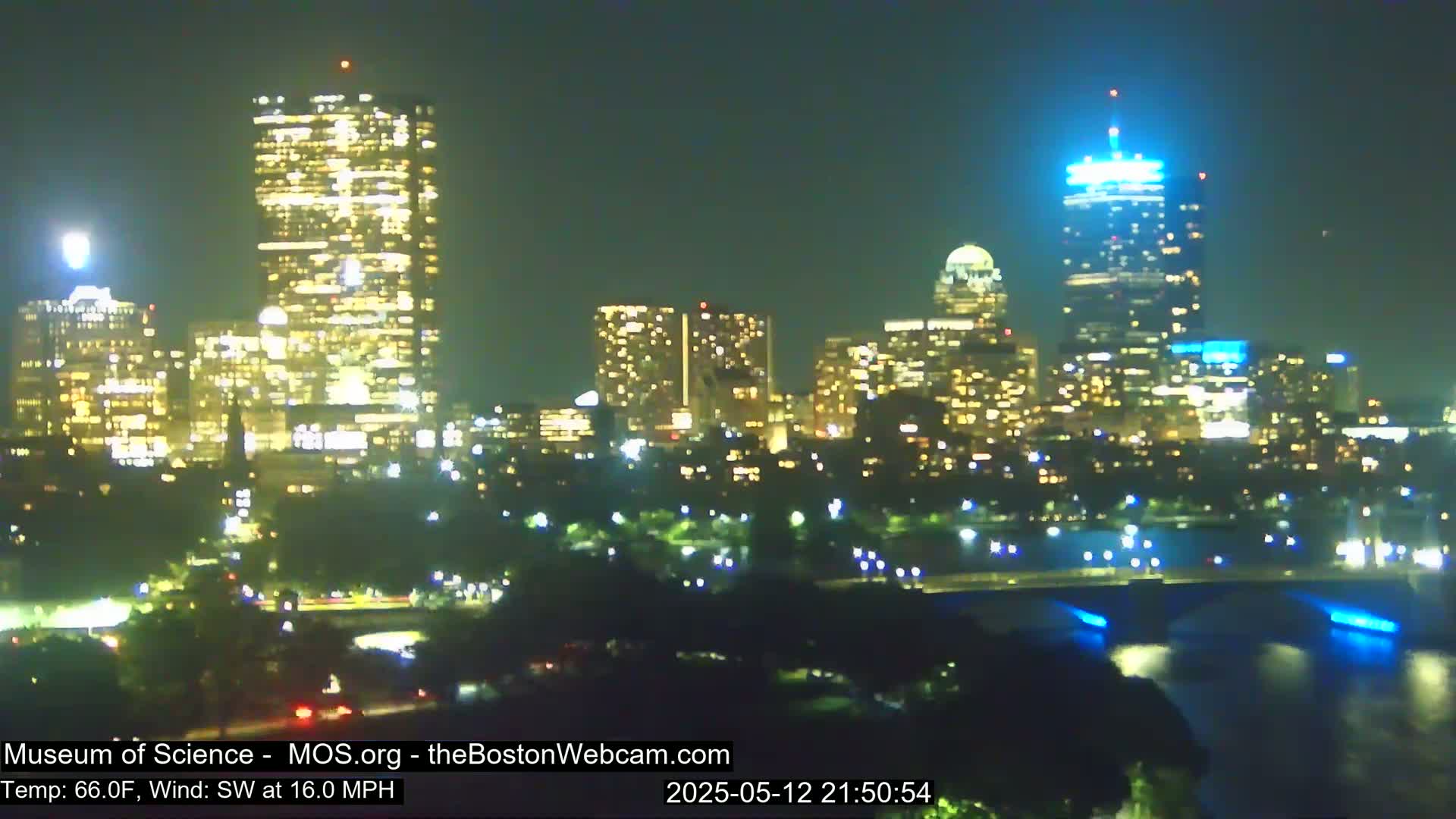 A nighttime view of a city skyline illuminated by lights, with a bridge and body of water in the foreground, under a clear sky and a southwest wind of 16 mph.