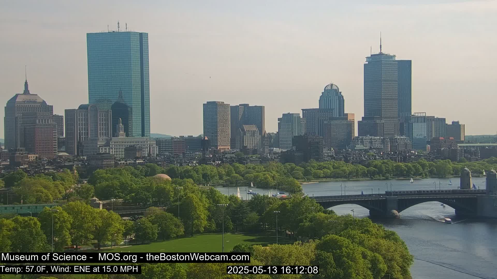 A city skyline with many tall buildings is visible across a river, with a bridge and green park in the foreground on a partly sunny day with a temperature of 57°F and an east-northeast wind at 15 mph.