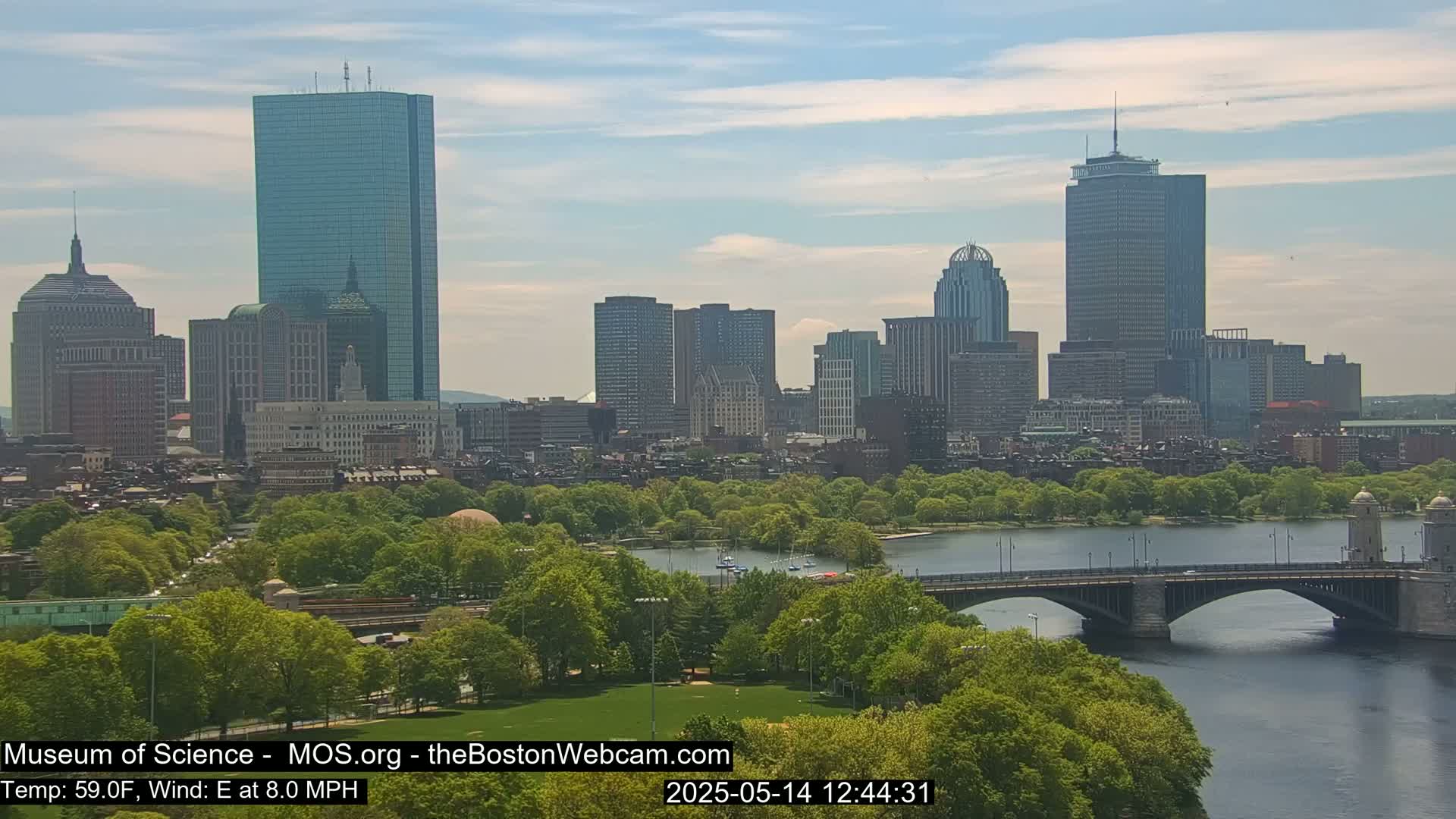A city skyline with numerous skyscrapers is visible across a river, under a partly cloudy sky; a bridge is in the foreground, and lush green trees line the riverbanks.