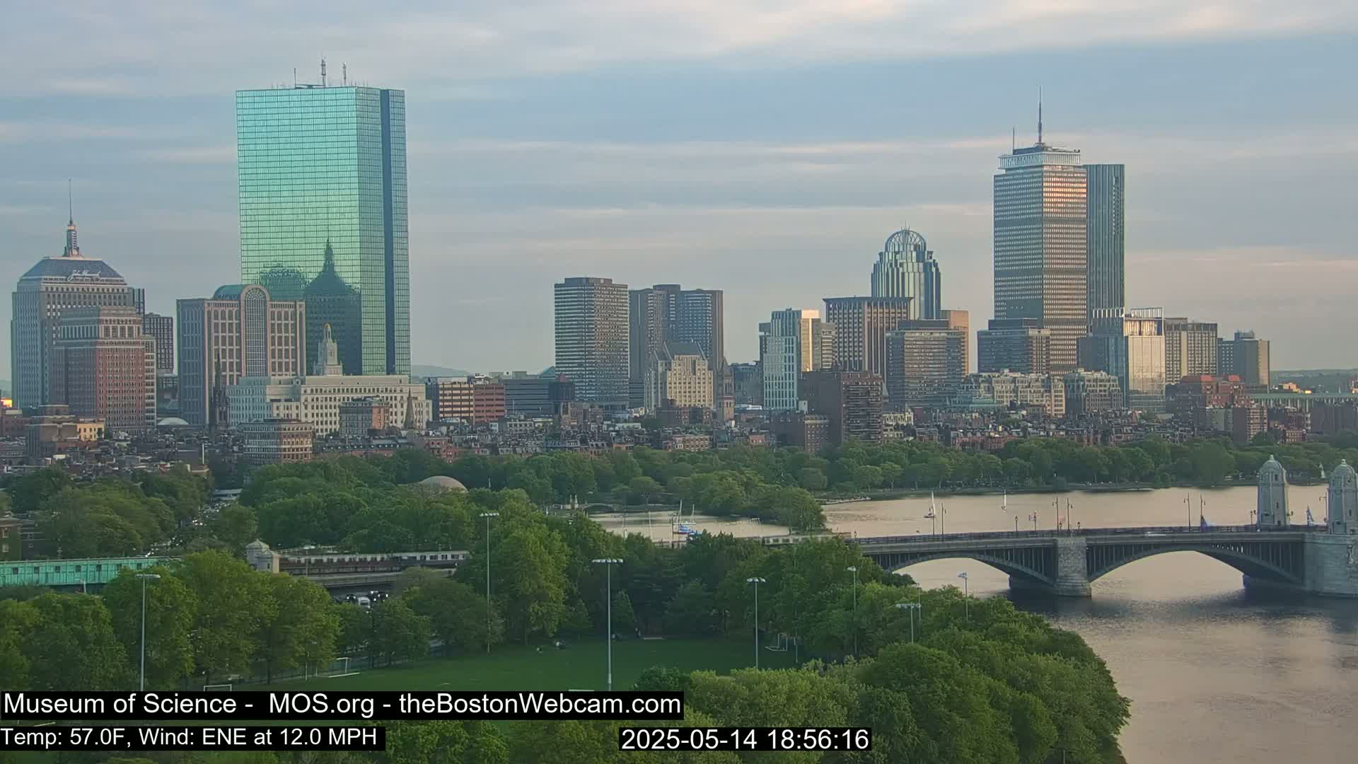 A city skyline with numerous skyscrapers and high-rise buildings is visible across a river, with a bridge and green parkland in the foreground, under a partly cloudy sky.