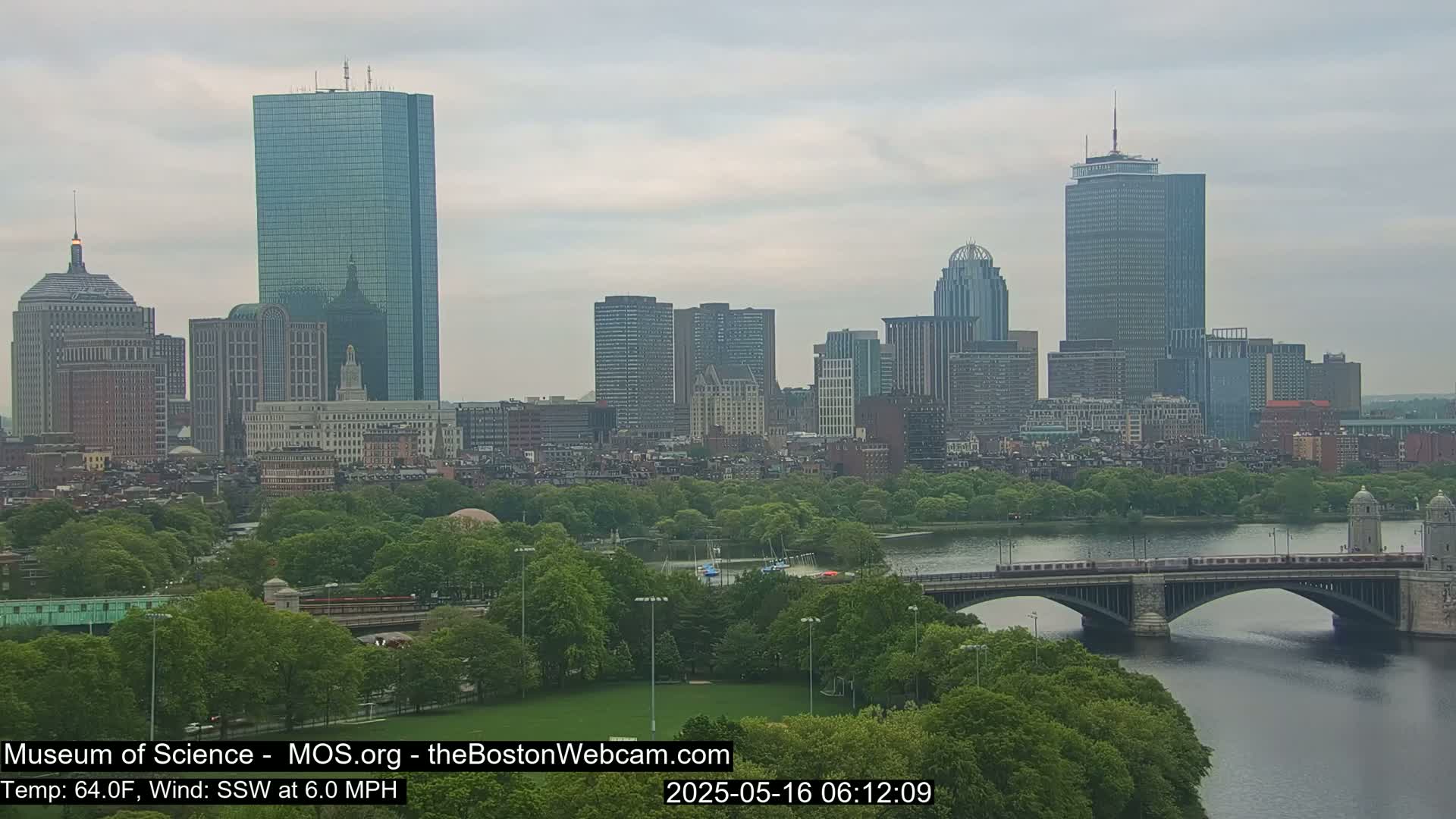 A city skyline with numerous skyscrapers and other buildings is visible across a river with a bridge, under an overcast sky.