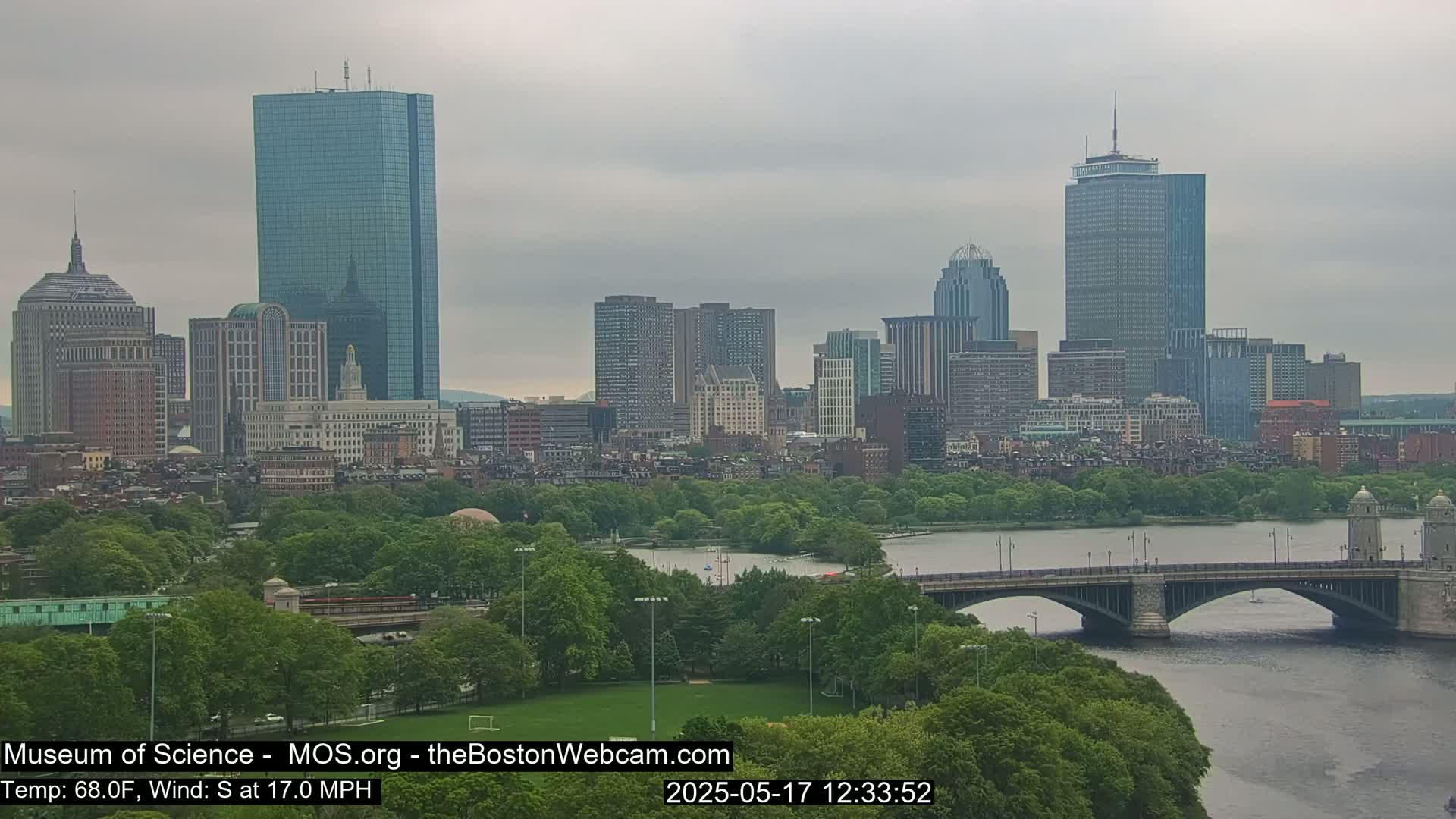 A city skyline with numerous skyscrapers is visible across a body of water, under an overcast sky, with a bridge in the foreground and lush green trees lining the riverbank.