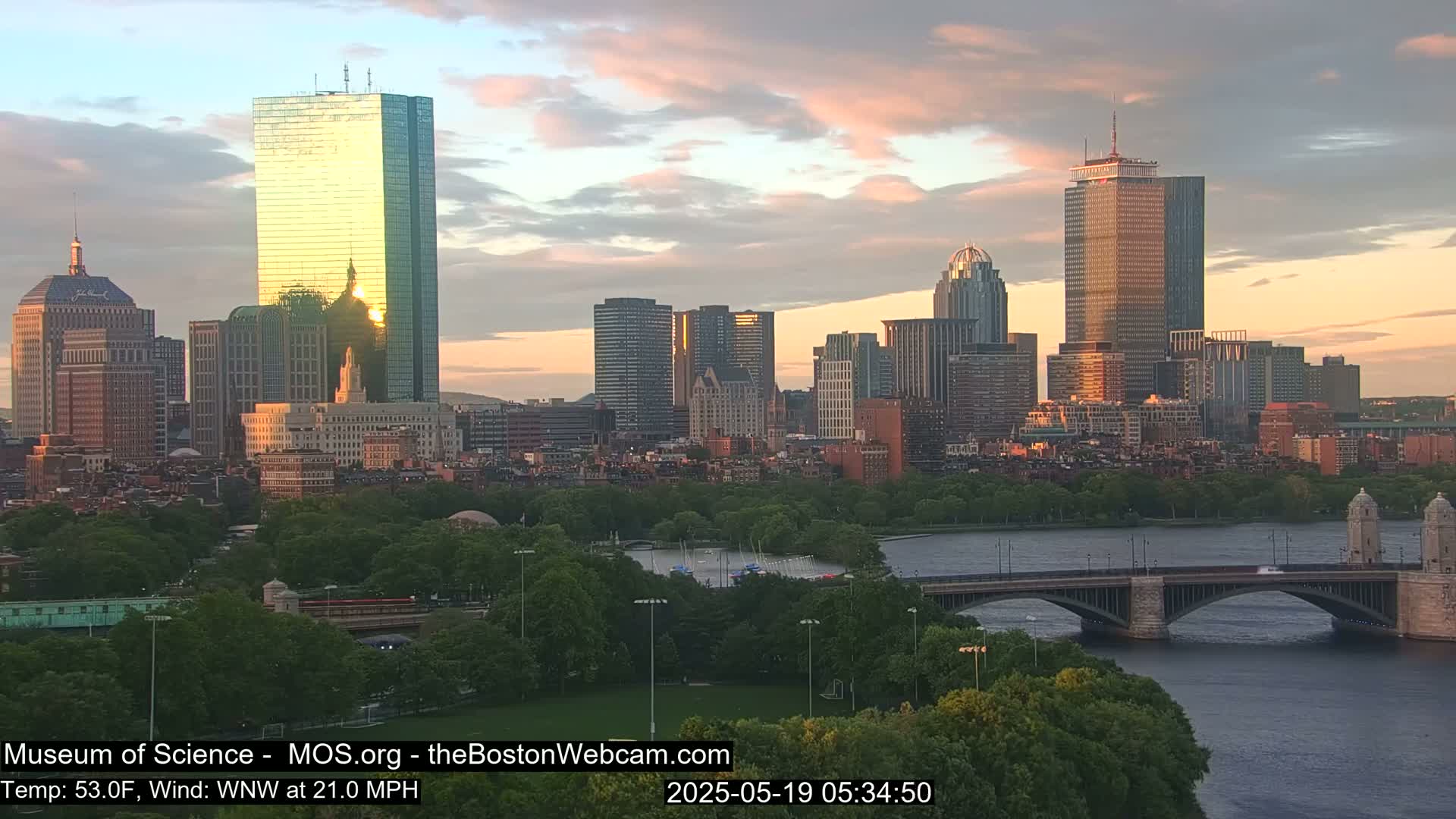 A cityscape at sunset with a bridge crossing a river, under partly cloudy skies.